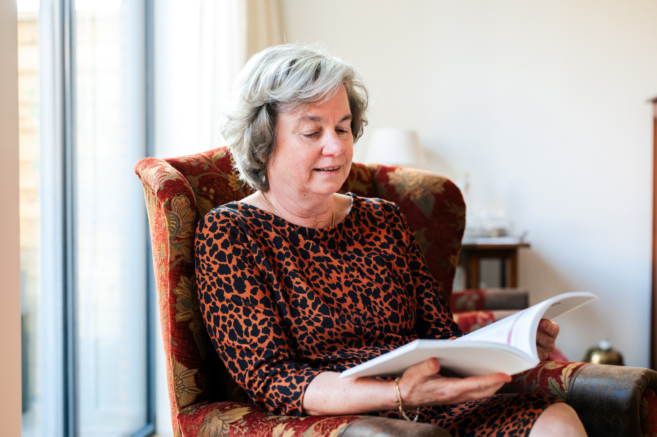 Vrouw met grijs haar en zwart met bruine jurk in een stoel, lezend in een boek.