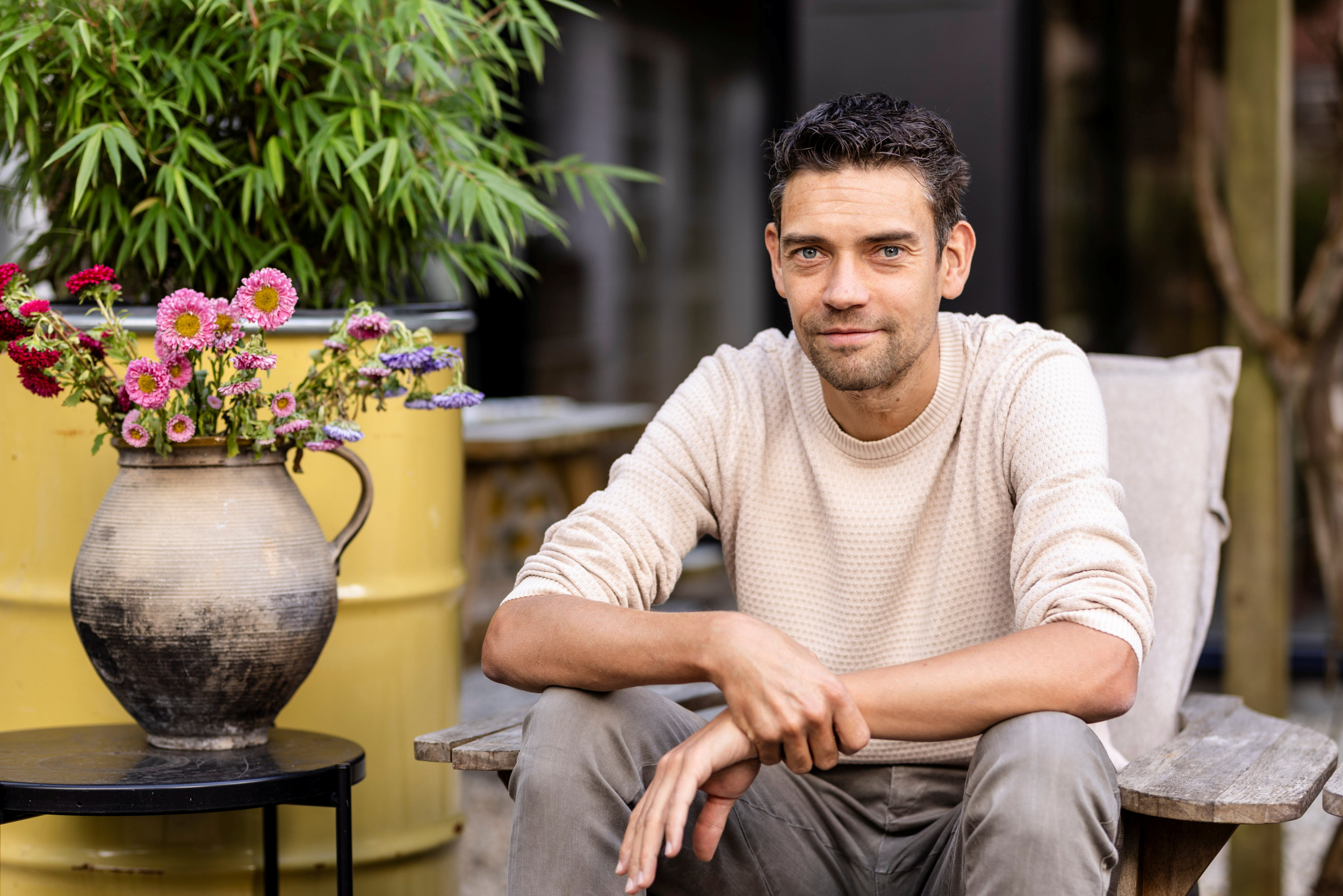 Jonge man met zwart haar en witte trui, zittend in een stoel naast een kan met bloemen.