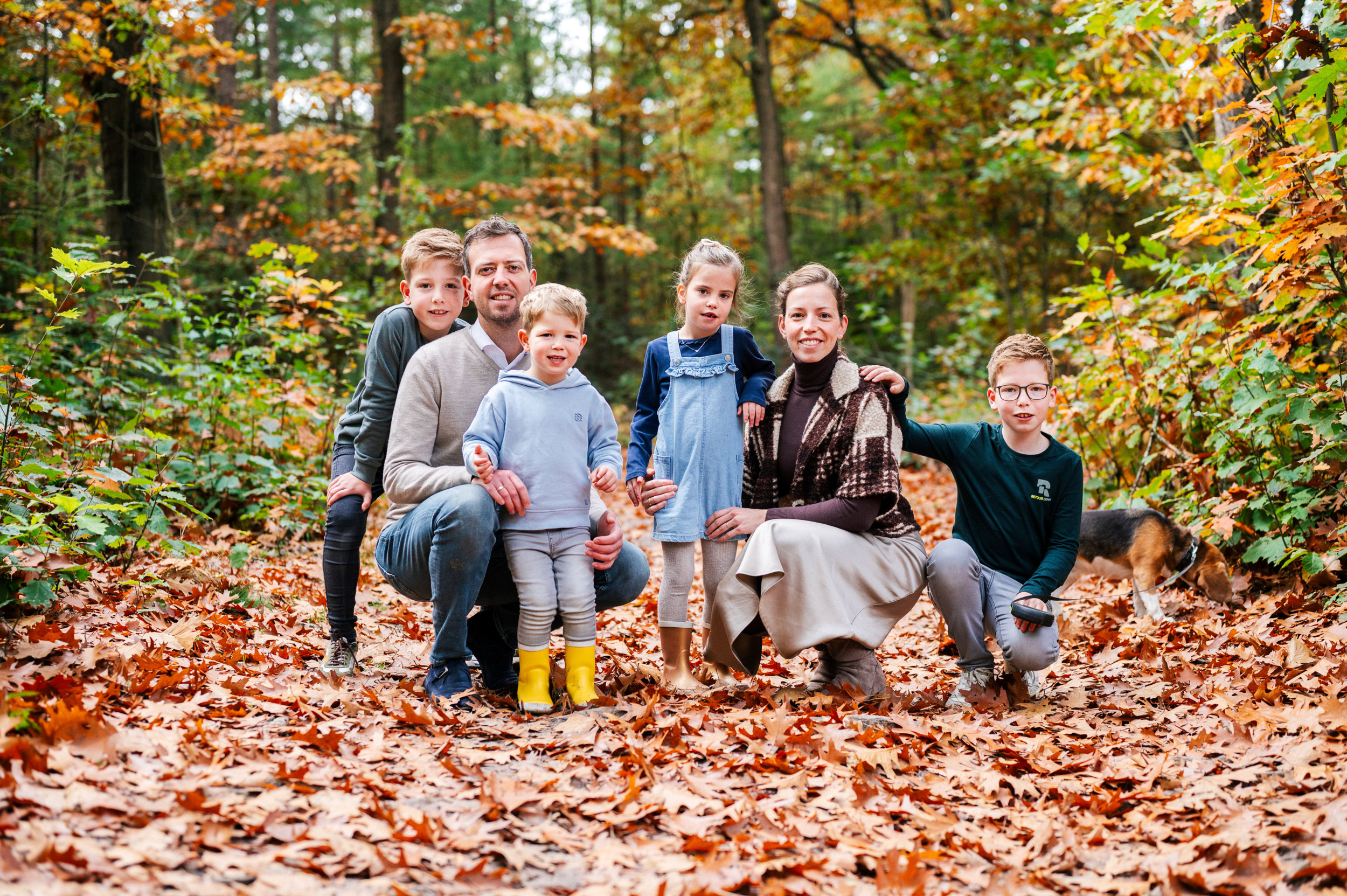 Een gezin in een herfstbos. Links een man in beige trui en jeans, op zijn hurken. Rechts een vrouw in beige rok, bruine coltrui en teddy vestje op haar hurken. Om hen heen vier jonge kinderen: drie jongens en een meisje.