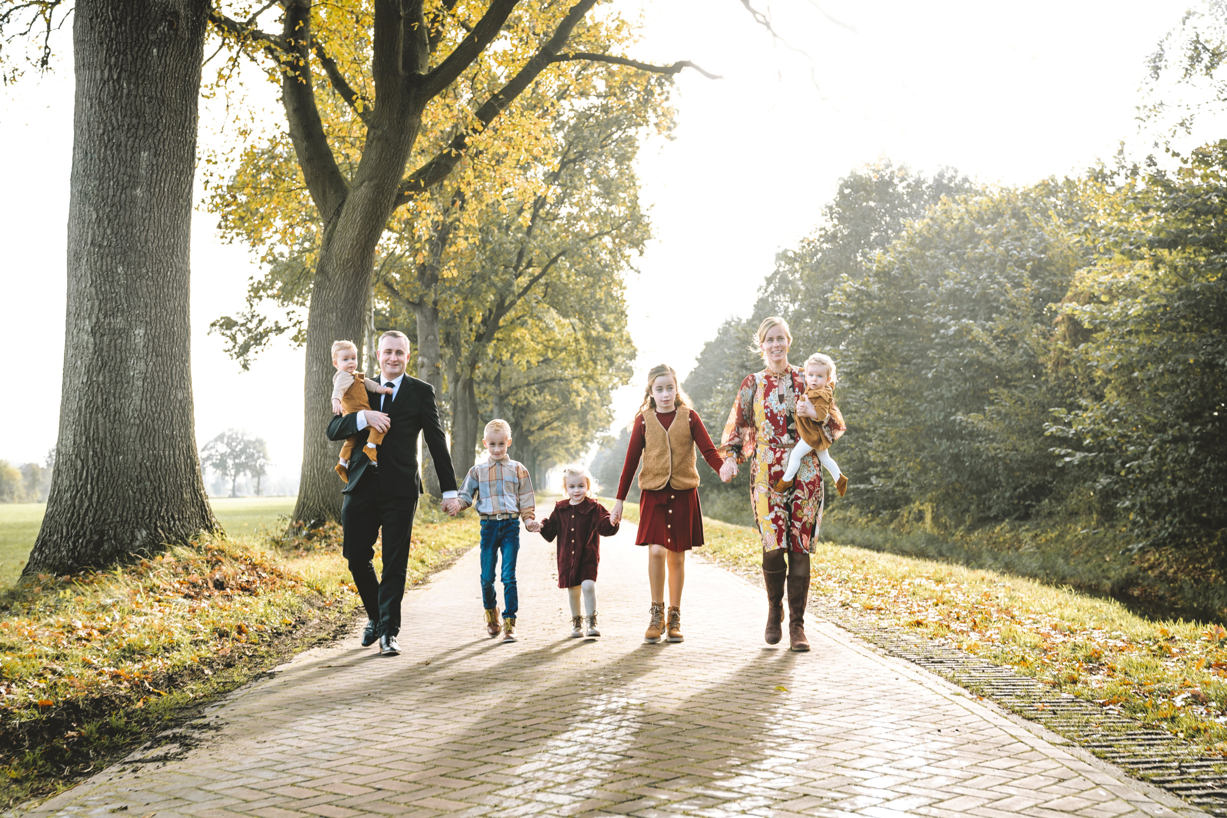 Man in zwart pak met jongetje op de arm en vrouw in kleurige jurk met meisje op de arm; ertussen drie oudere kinderen. Hand in hand op een landweg.