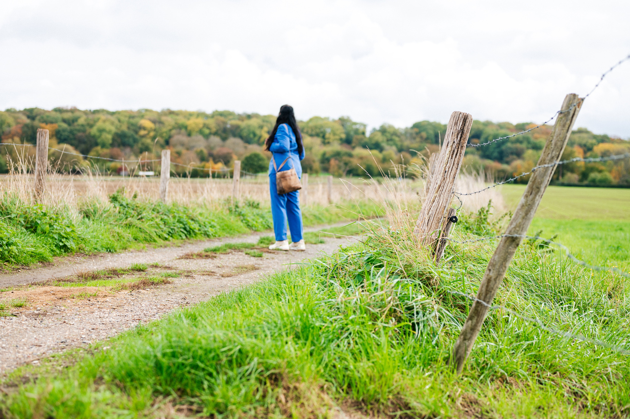 Vrouw in blauw broekpak, onherkenbaar gefotografeerd, in een landelijke omgeving met bos op de achtergrond.