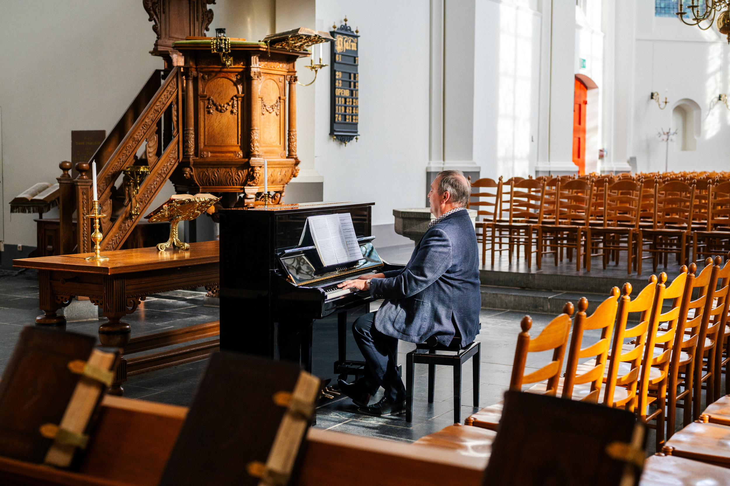 Een man in een blauw pak zit in een oude kerk bij een oude preekstoel piano te spelen.