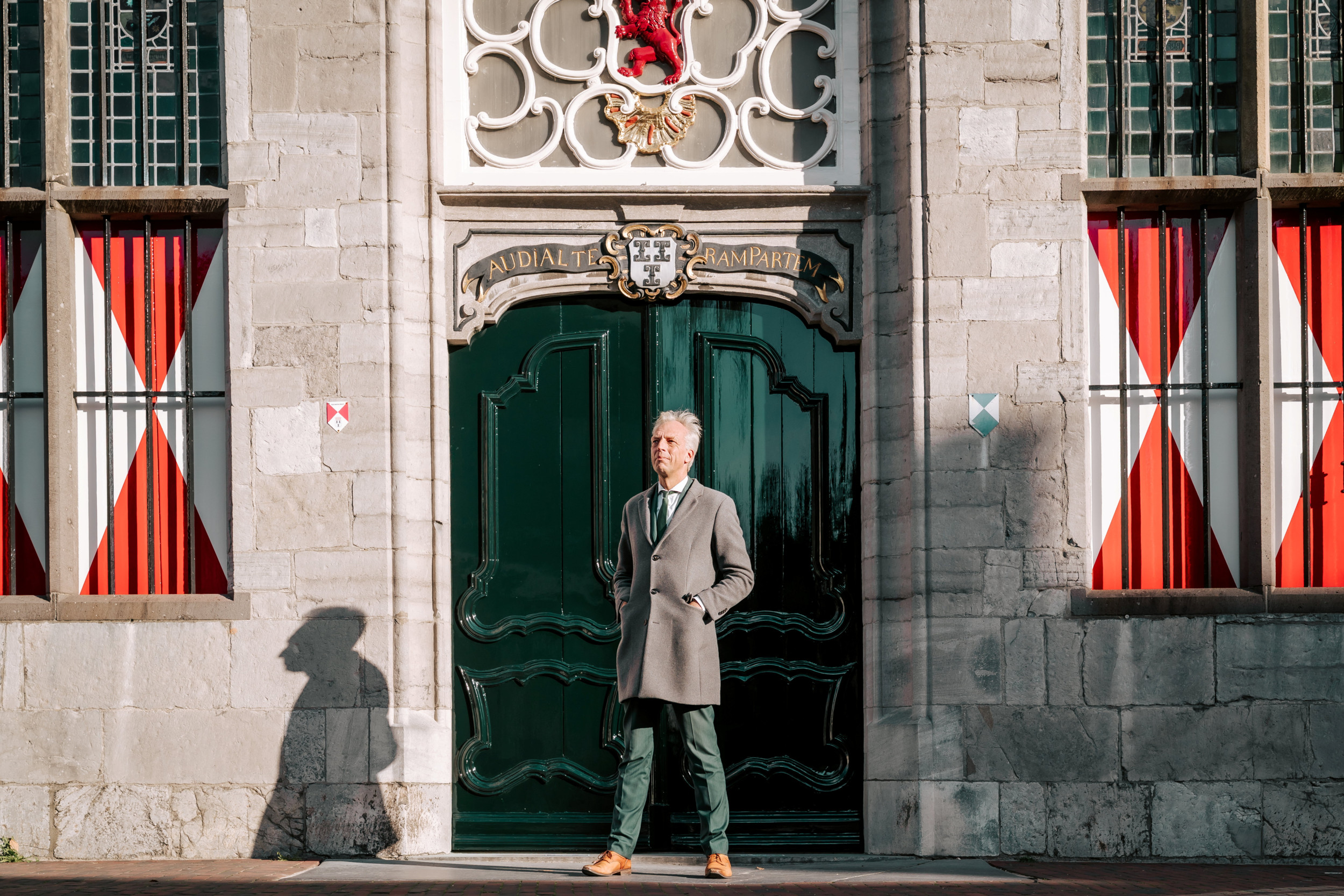 Man met grijs haar, groen kostuum en lichte overjas voor de deur van een monumentaal gebouw,