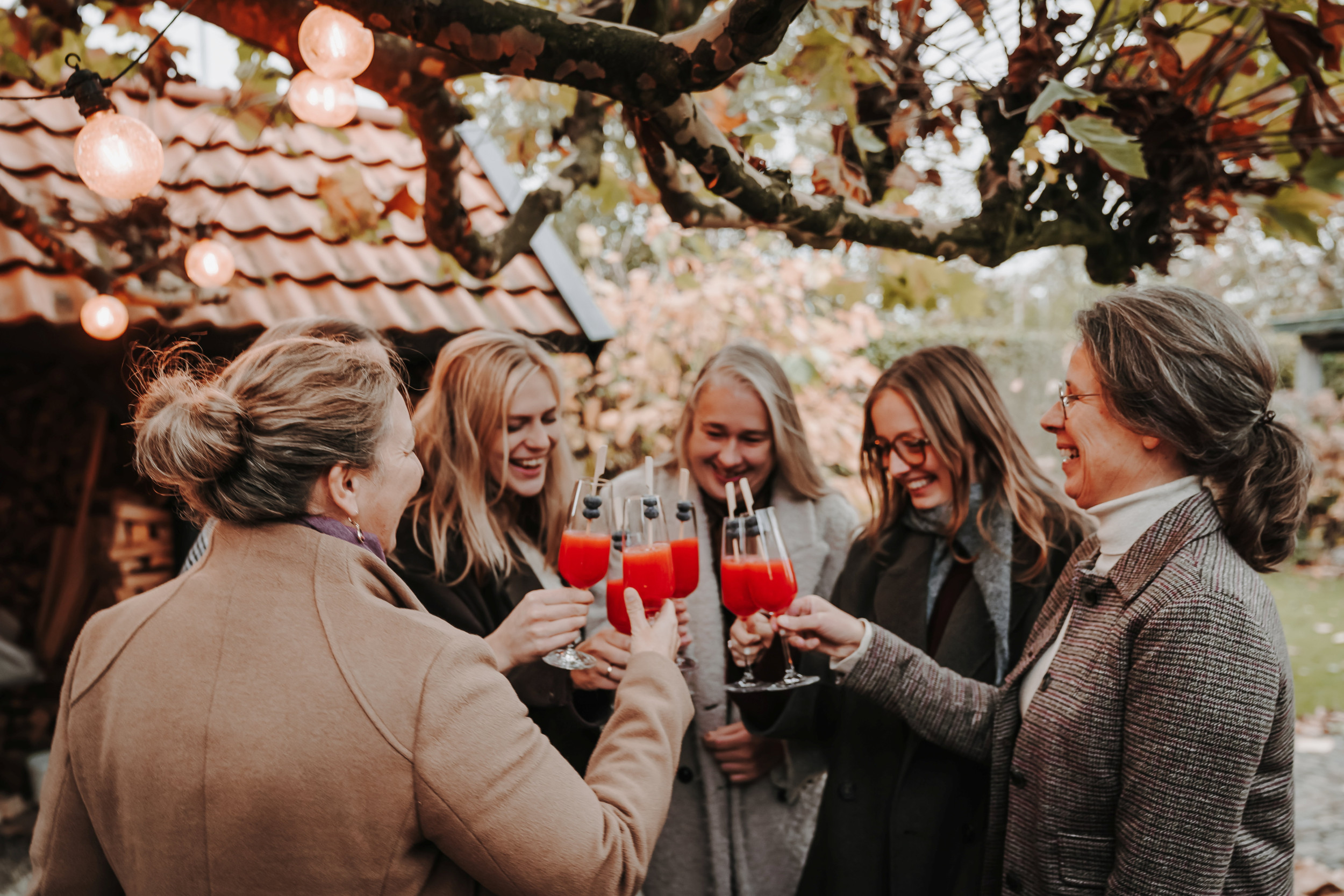 Vijf lachende vrouwen heffen champagneglazen met daarin rood sap, in een tuin met op de achtergrond een schuur.