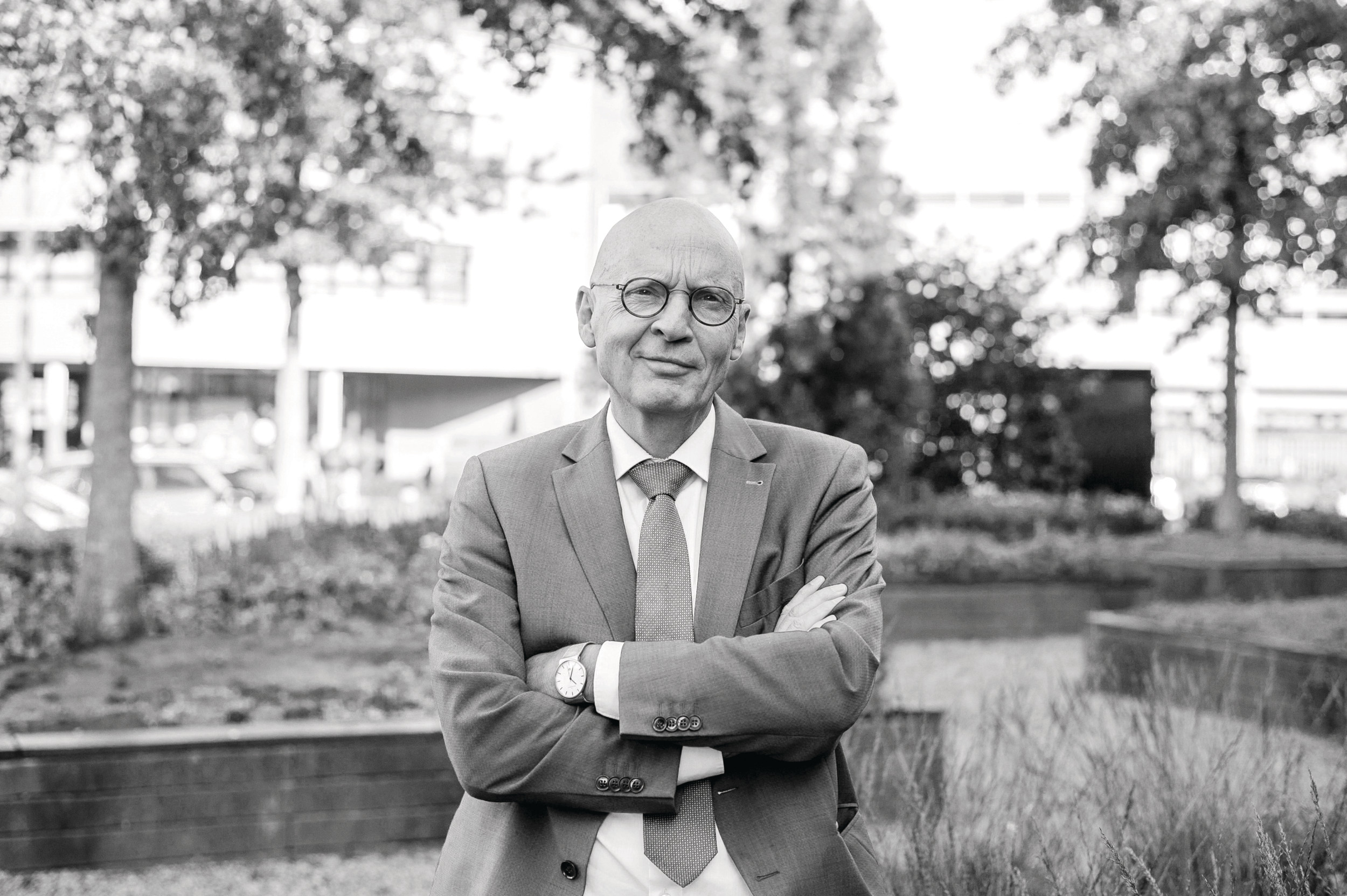 Portret van een man in pak, in een tuin bij een schoolgebouw.