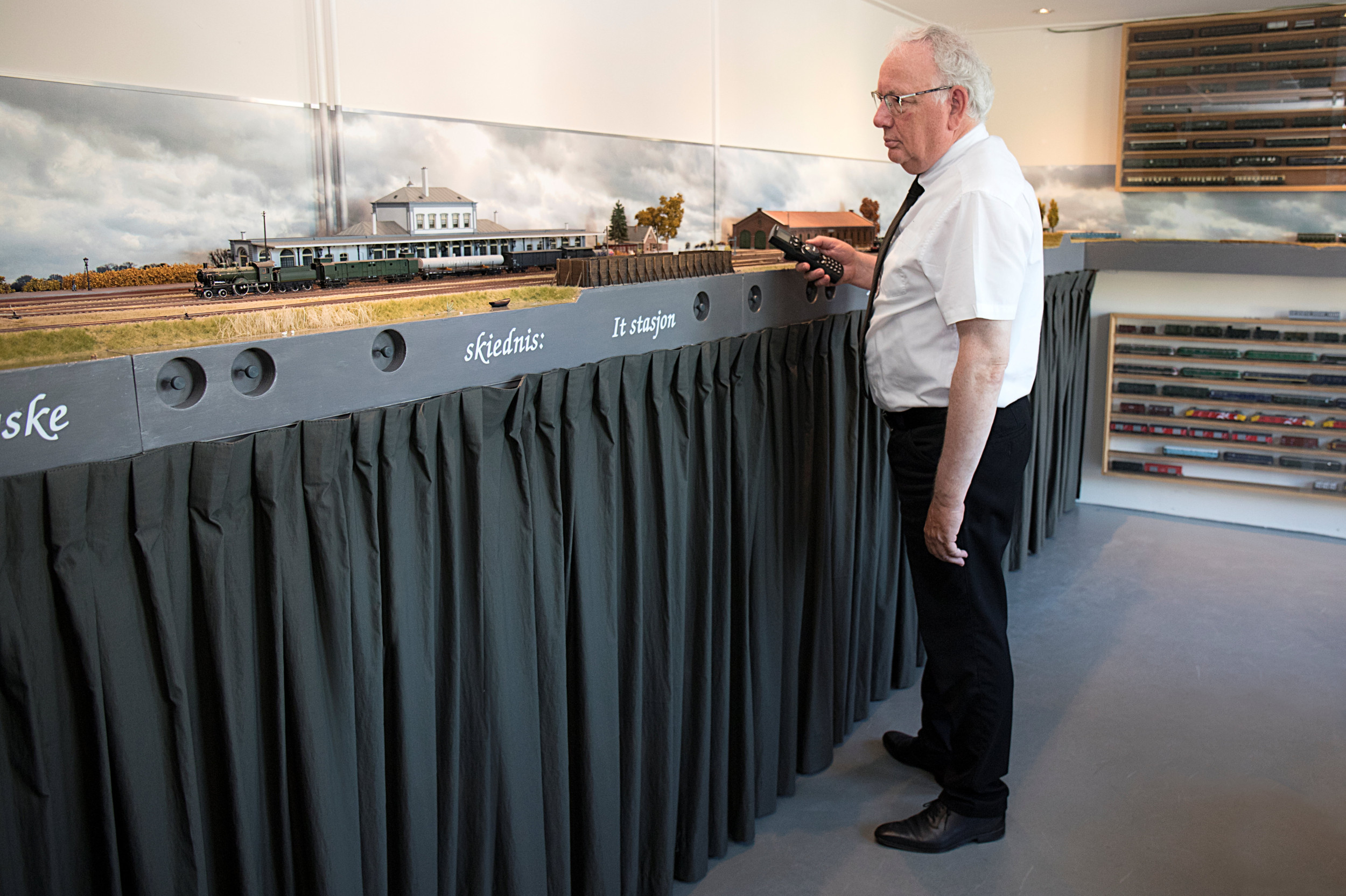 Een man in zwarte broek en wit overhemd staat bij een modelspoorbaan.