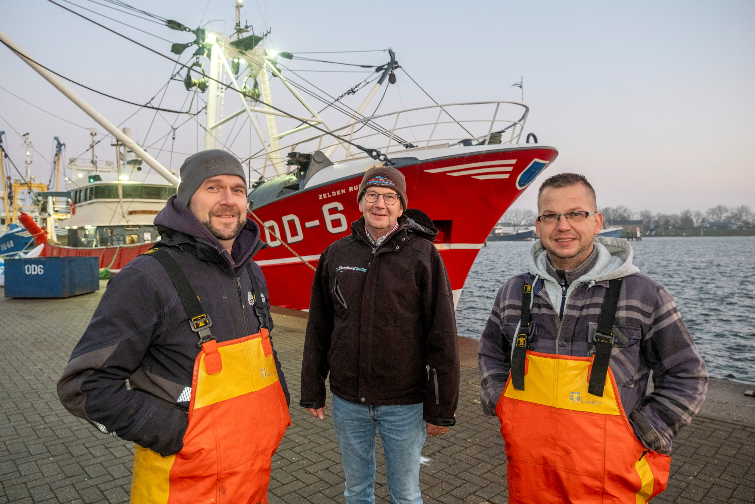 Drie mannen in werkkleding staan op de kade voor een rode viskotter met de naam Zeldenrust.