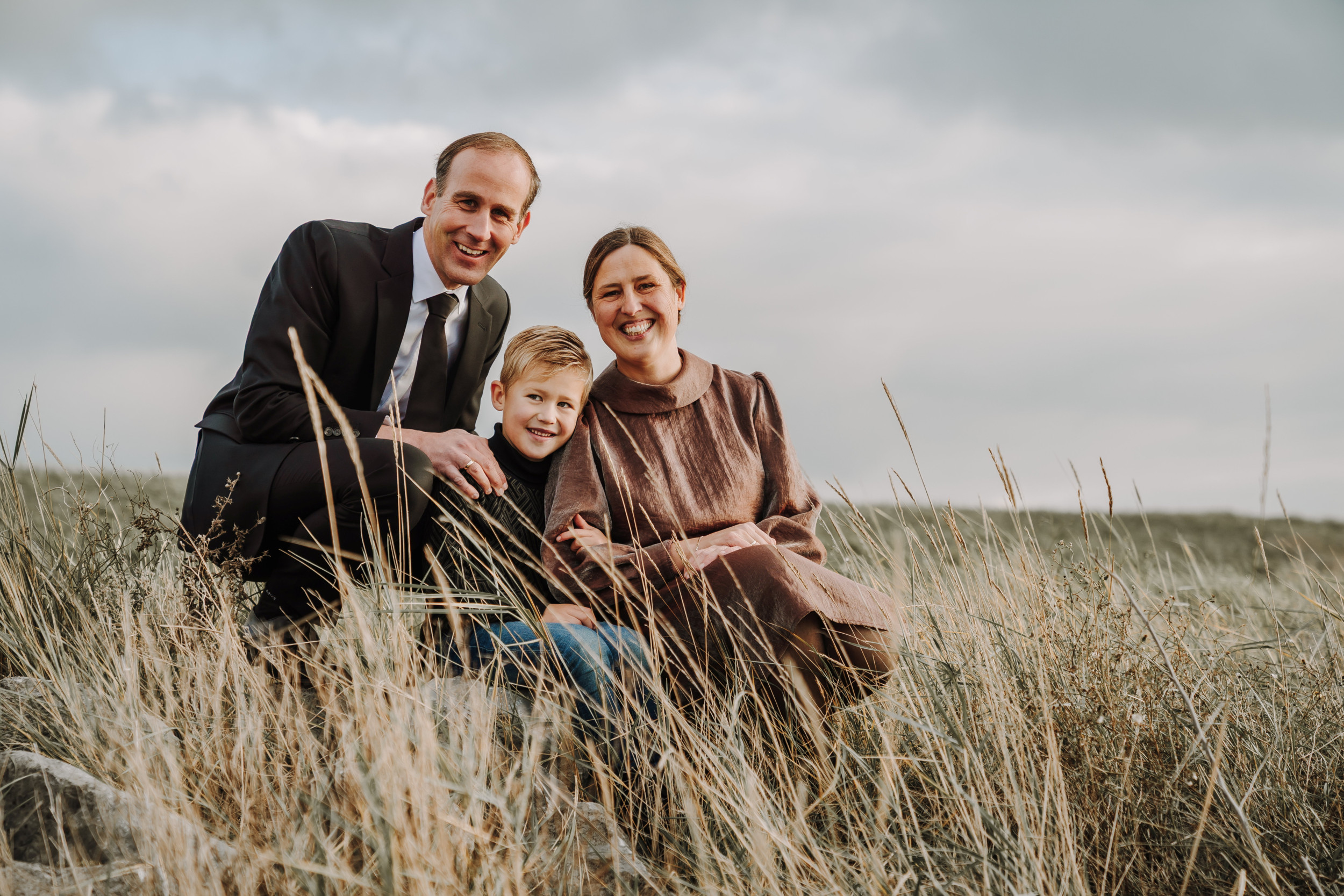 Man in donker pak met vrouw in een bruine jurk en tussen hen een blonde jongen in het gras. Op de achtergrond een grijze lucht.