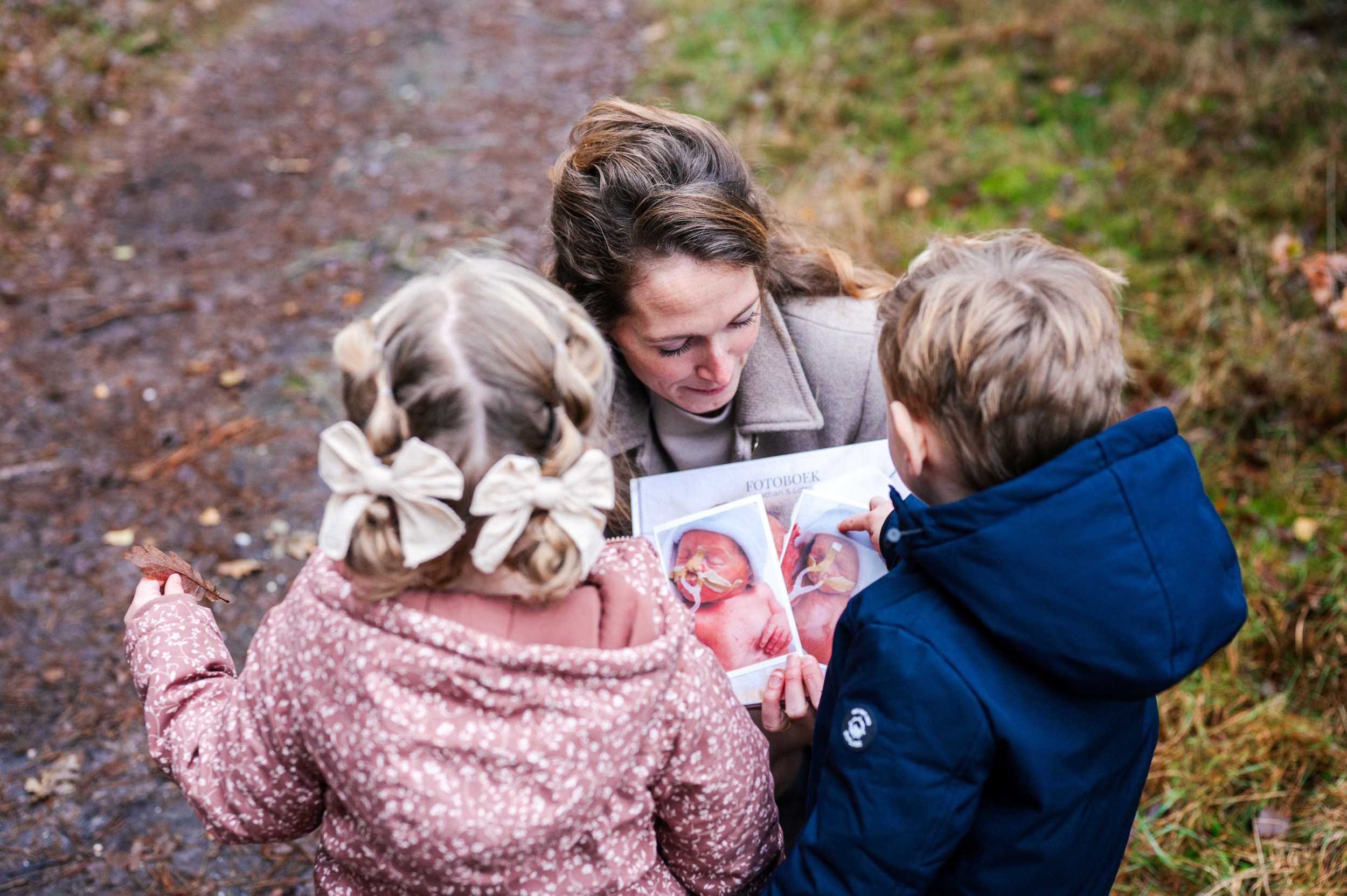 Jonge vrouw die op een bospad twee kleine kinderen een fotoboek laat zien met voorop de afbeeldingen van twee couveusekinderen.