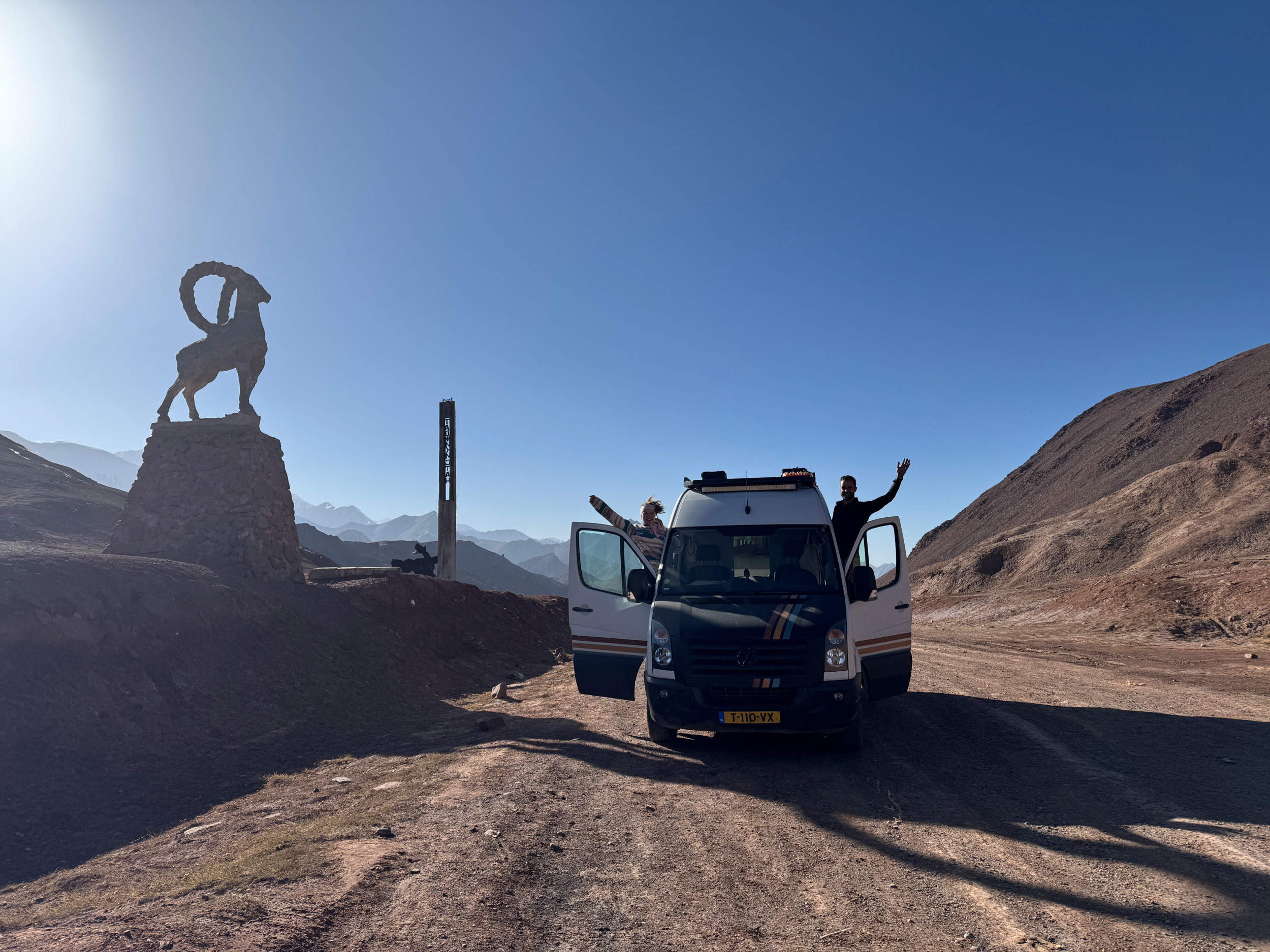Camper op een onverharde weg tussen bergen. Een man en een vrouw zwaaien vanuit de geopende portieren.