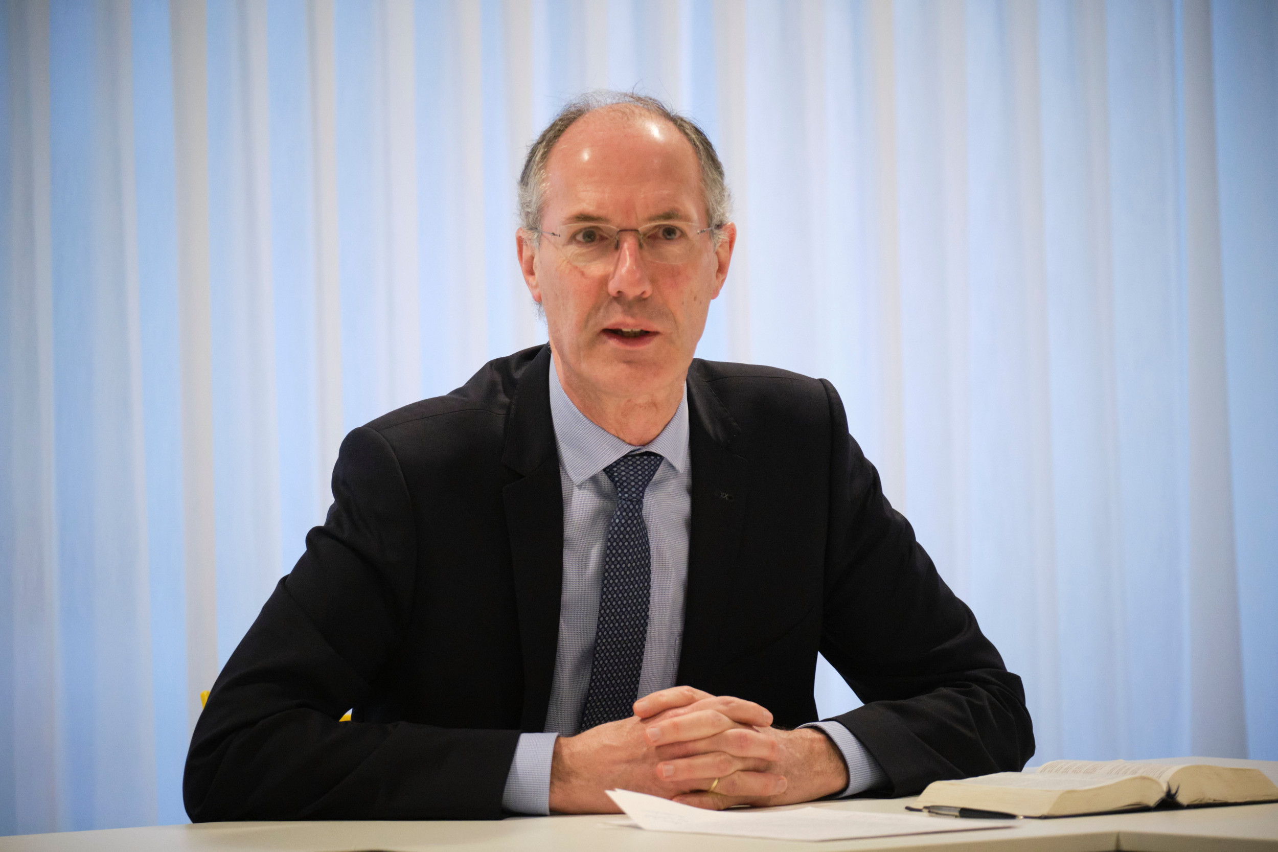 Man met dun grijs haar en blauw kostuum, zittend achter een tafel met gevouwen handen.