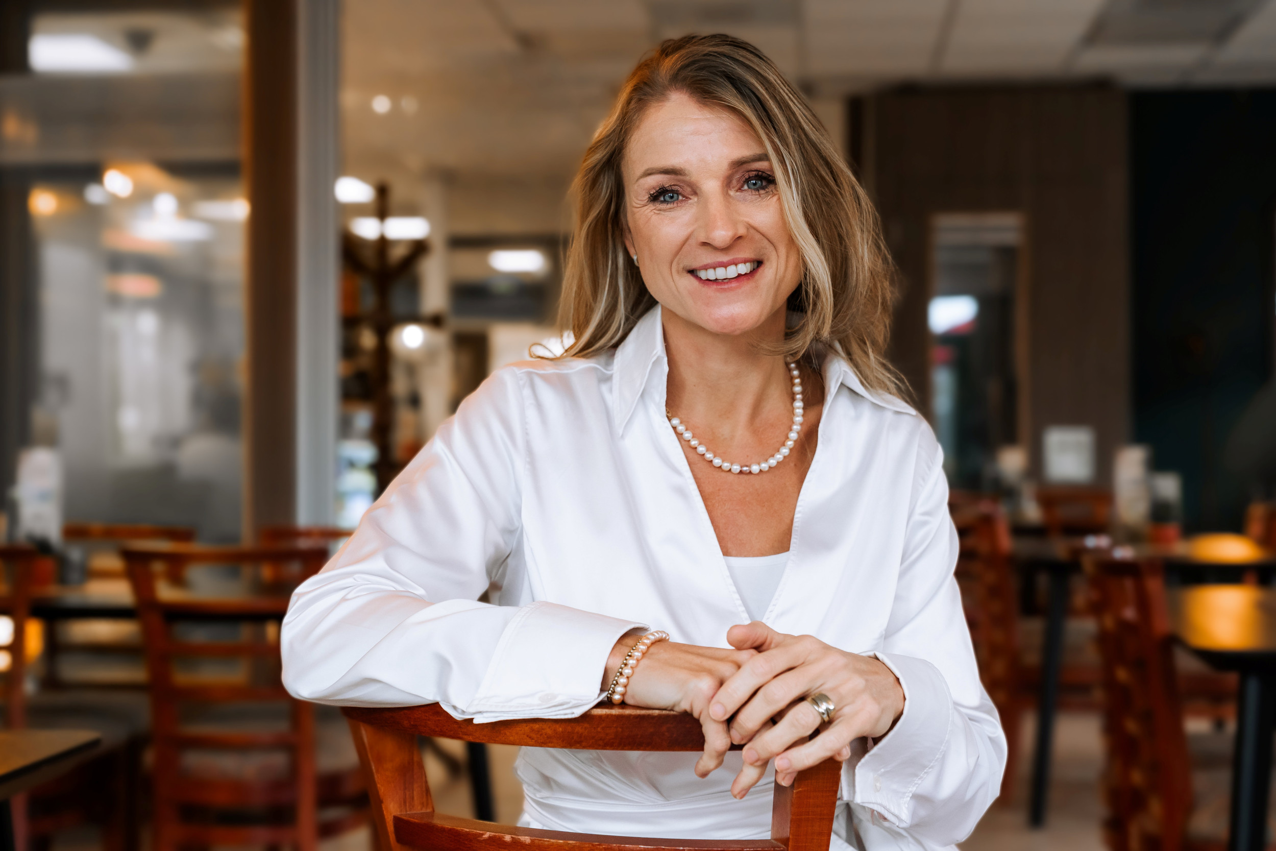 Burgemeester de Jonge, lang blond haar, witte blouse en parelketting, zit op een stoel in een Nijkerks ontmoetingscentrum.