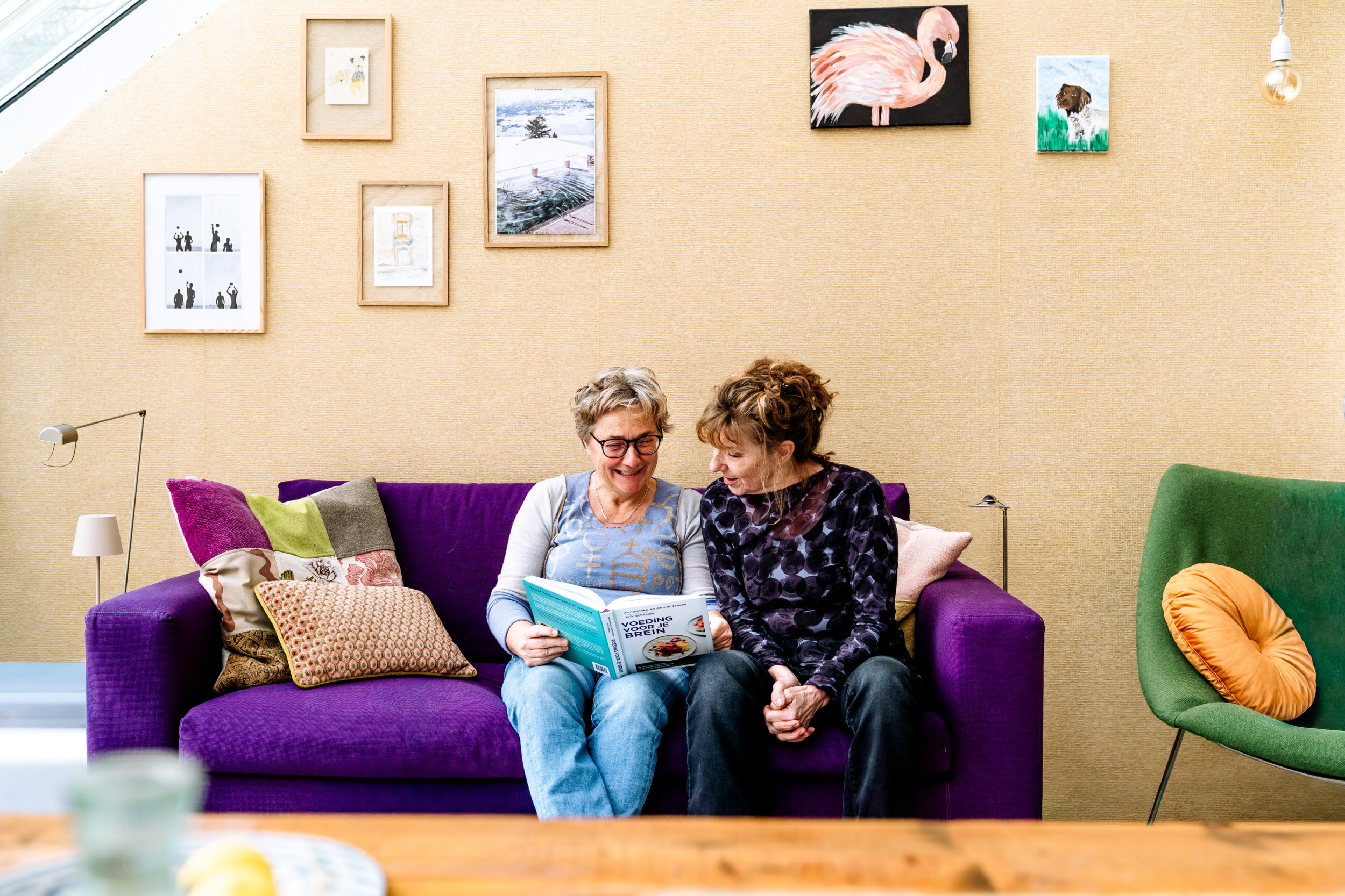Twee vrouwen van middelbare leeftijd naast elkaar op een paarse bank, kijkend in een boek dat een van de vrouwen in de handen heeft.