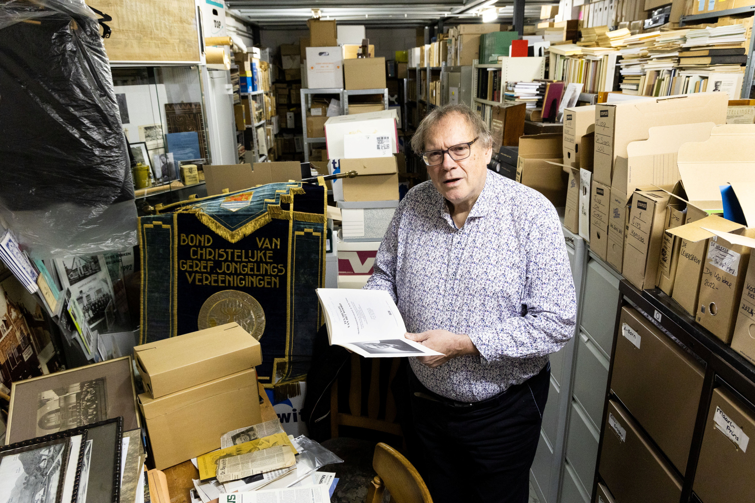 Man van middelbare leeftijd met een boek in de hand in een archiefruimte vol kasten, stellingen en dozen.