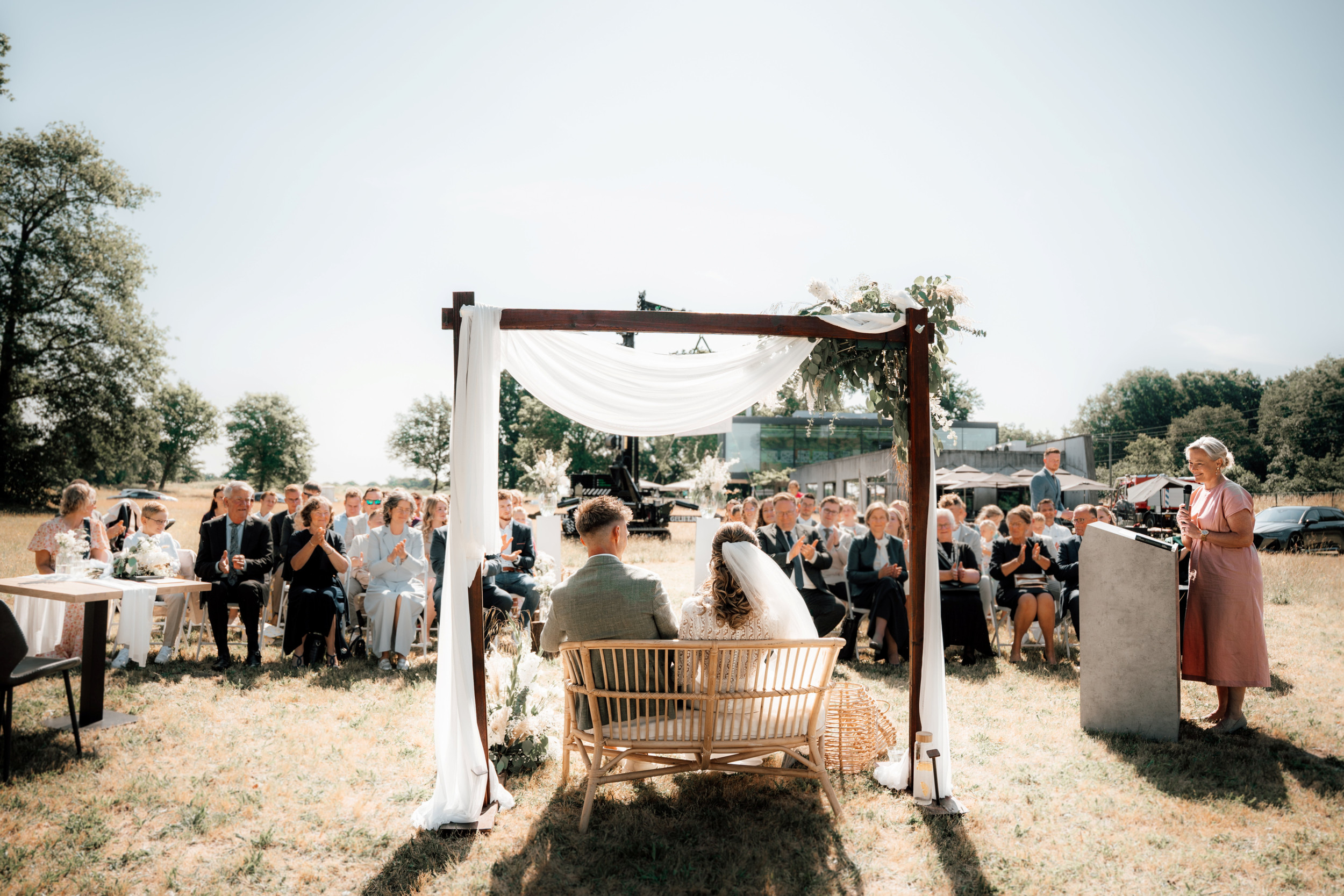 Vrouw in een roze jurk achter een katheder die in de openlucht een bruidspaar toespreekt.
