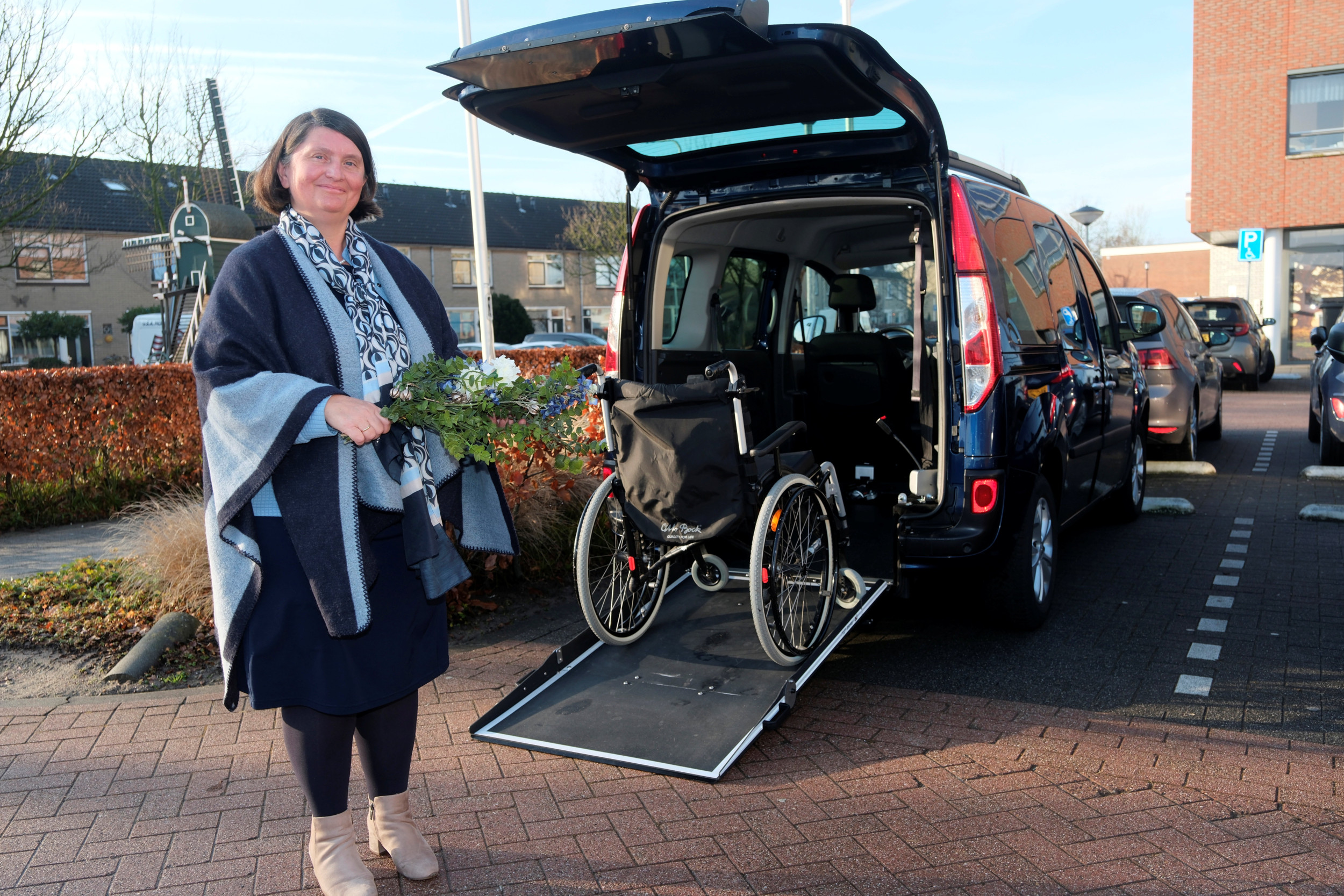 Vrouw met bruin boblijn-kapsel, donkerblauwe kleding en donkerblauwe omslagdoek met een boeket in de hand. Op de achtergrond een donkere bus met de klep open en een rolstoel op de rijplaat.