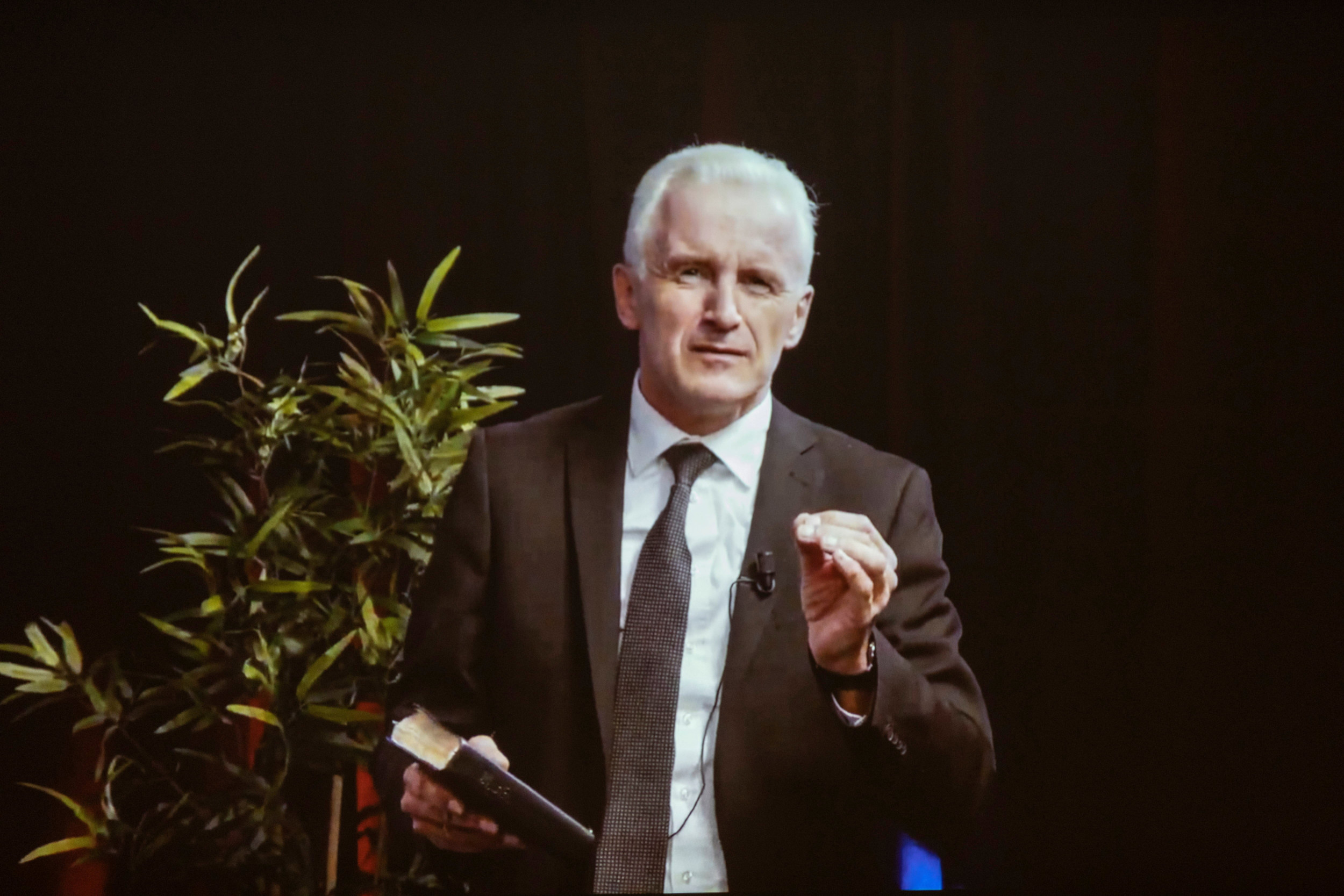 Man van middelbare leeftijd in donker pak, sprekend met opgeheven hand, in de andere hand een Bijbel.