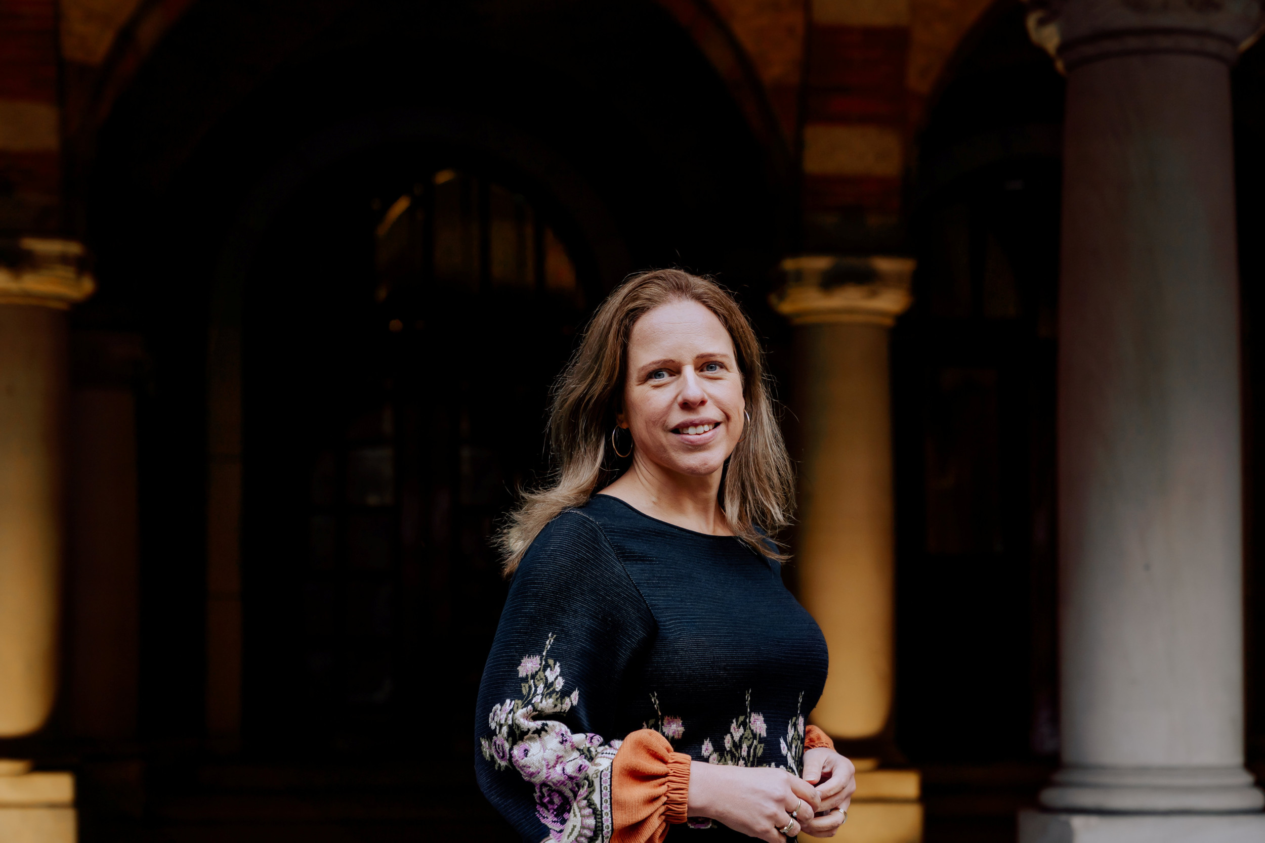 Burgemeesster Carola Schouten staat naast een pilaar in het gemeentehuis van Rotterdam. Ze heeft lang, blond haar en een zwarte gebloemde jurk aan.