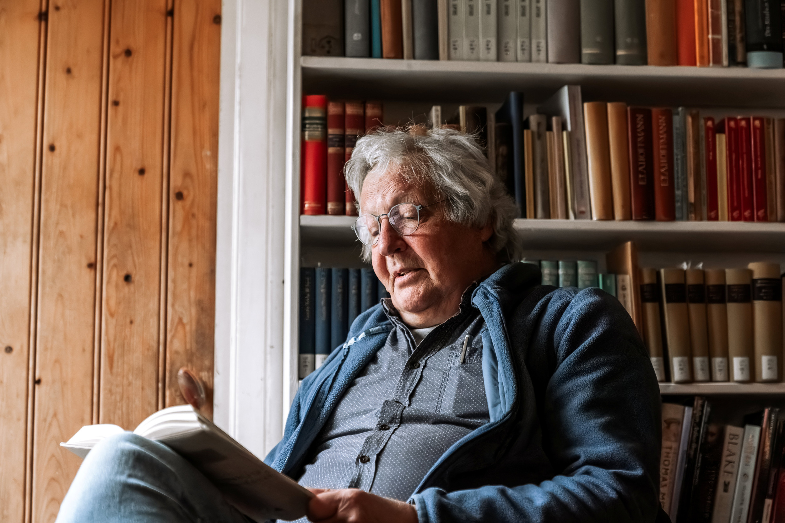 Een man met grijs haar leest in een boek. Hij zit voor een boekenkast.