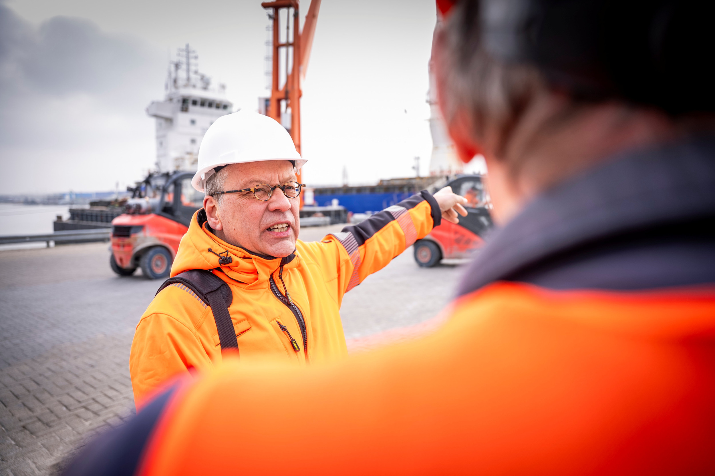Man in oranje jas en witte helm op het hoofd in gesprek op een kade met op de achtergrond een zeeschip.