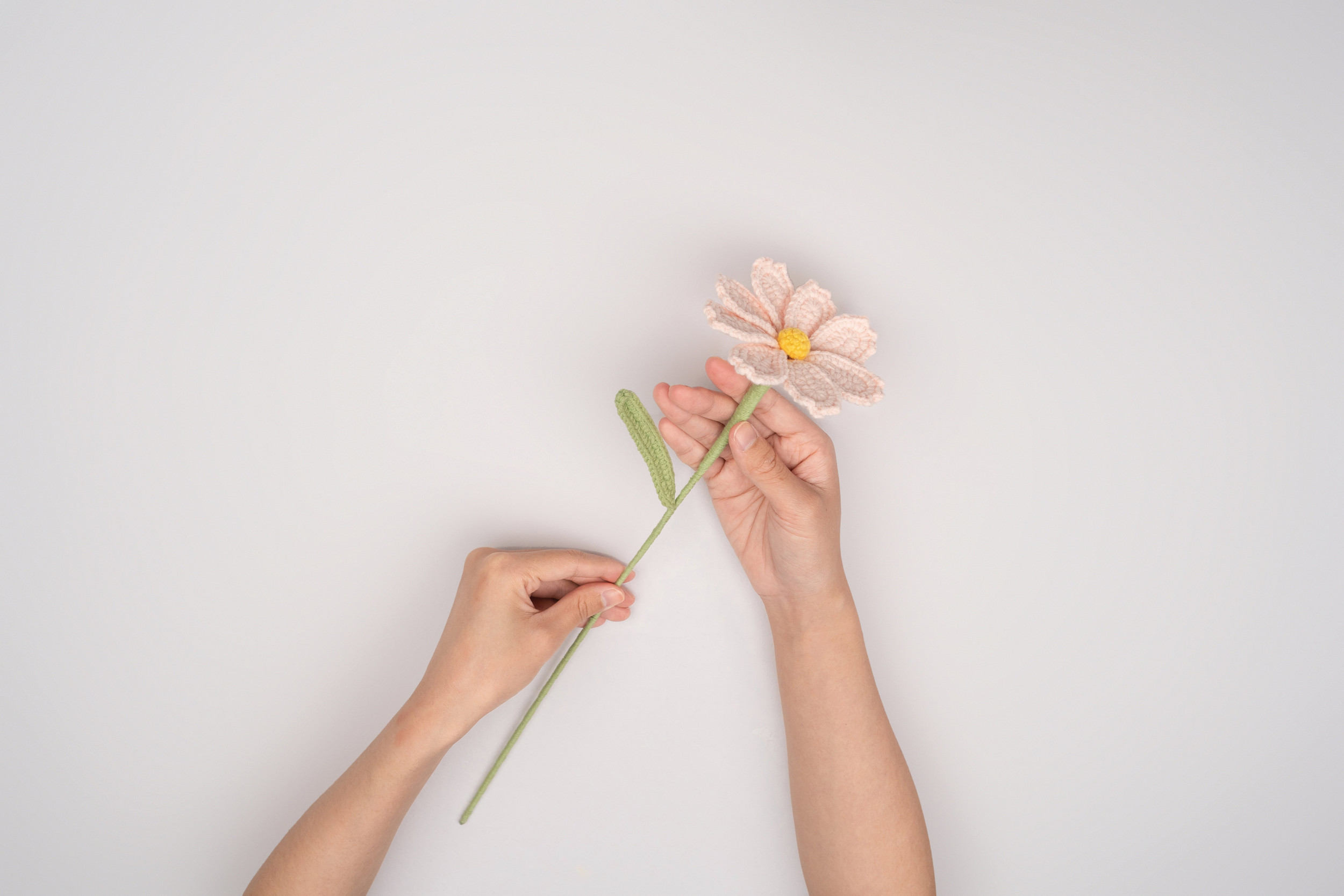 Twee handen die een zachtroze, gehaakte bloem vasthouden. De bloem heeft een lange, lichtgroene steel en een geel hart. 