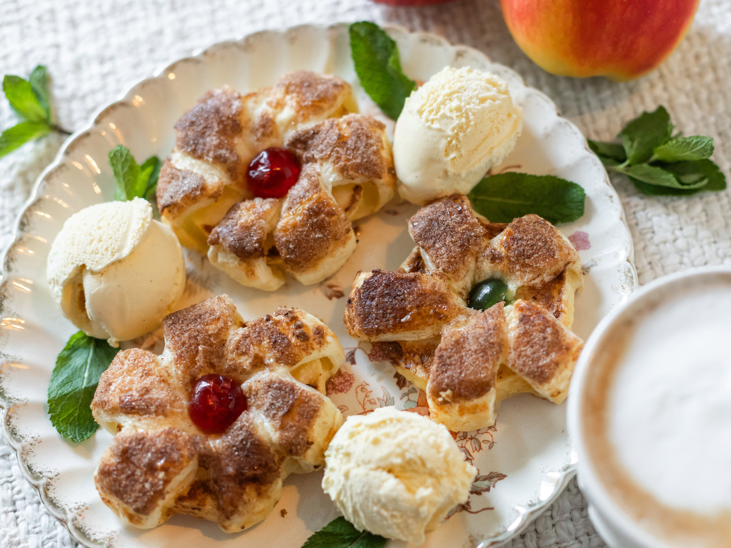Wit bord met daarom drie bolletjes ijs, gedecoreerd met muntblaadjes, en drie bloemvormige bladerdeegpakketjes met een geconfijte kers als bloemhart.