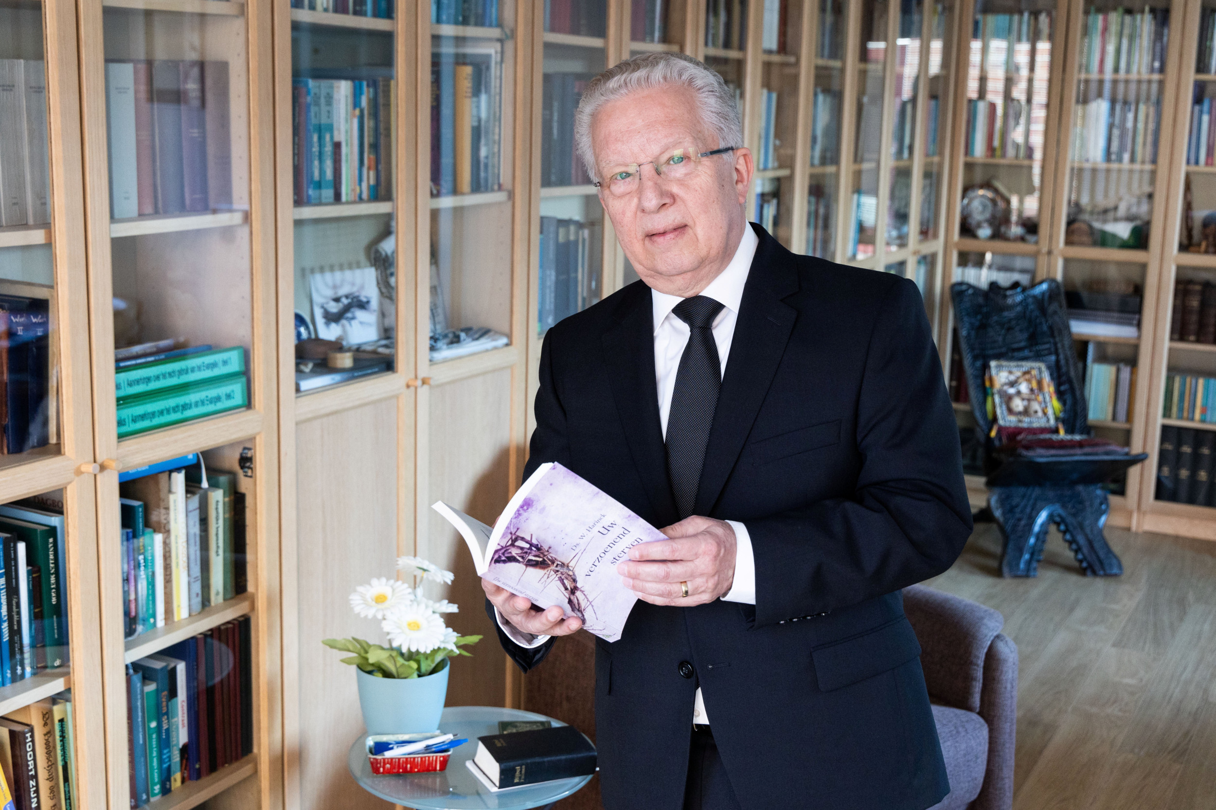 Man met bril, grijs haar en donker pak voor een boekenkast met een boekje in de hand.