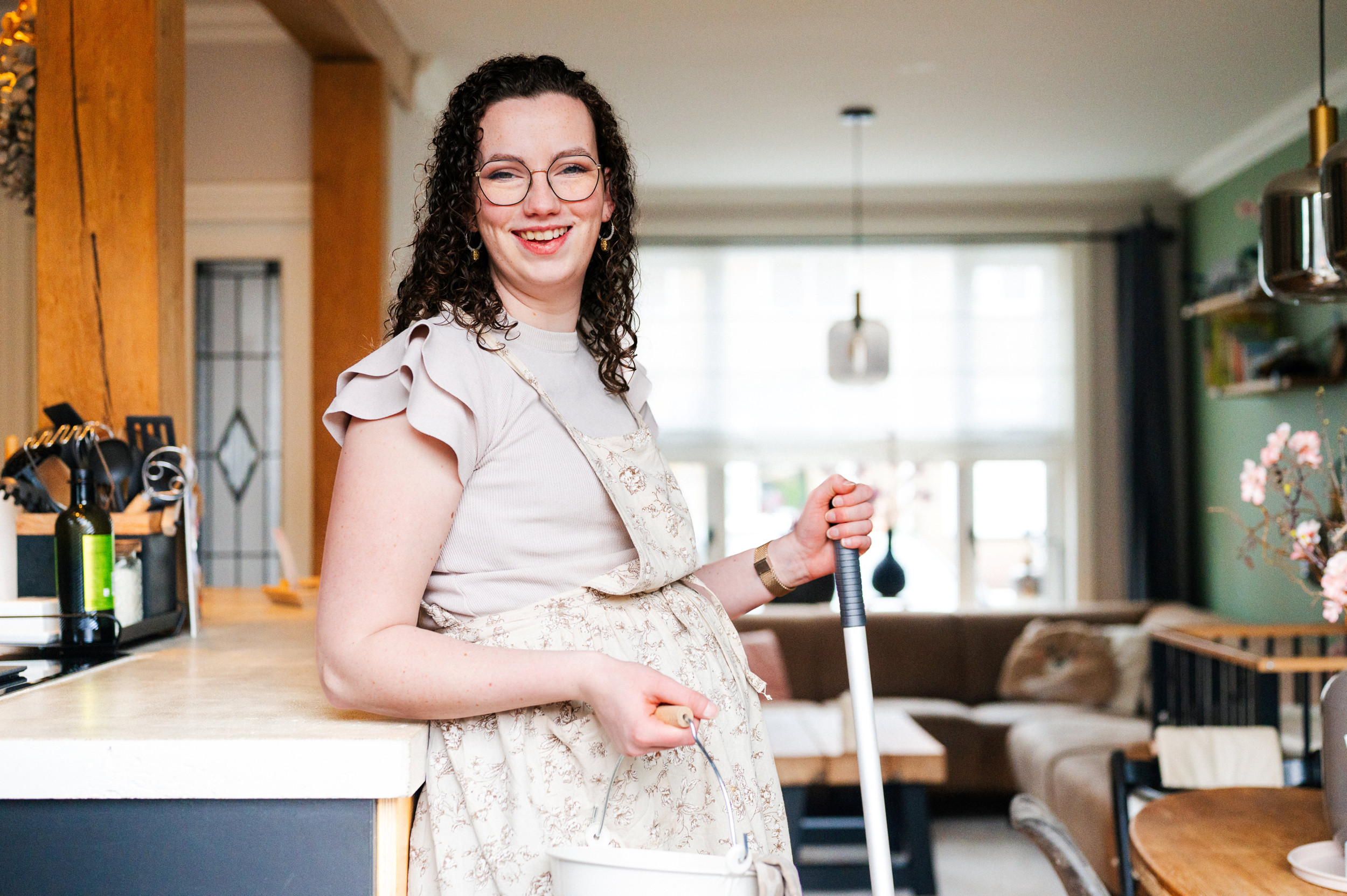Aline van den Boogaard (donkere krullen, bril, beige kleding) heeft een dweilstok in haar handen.