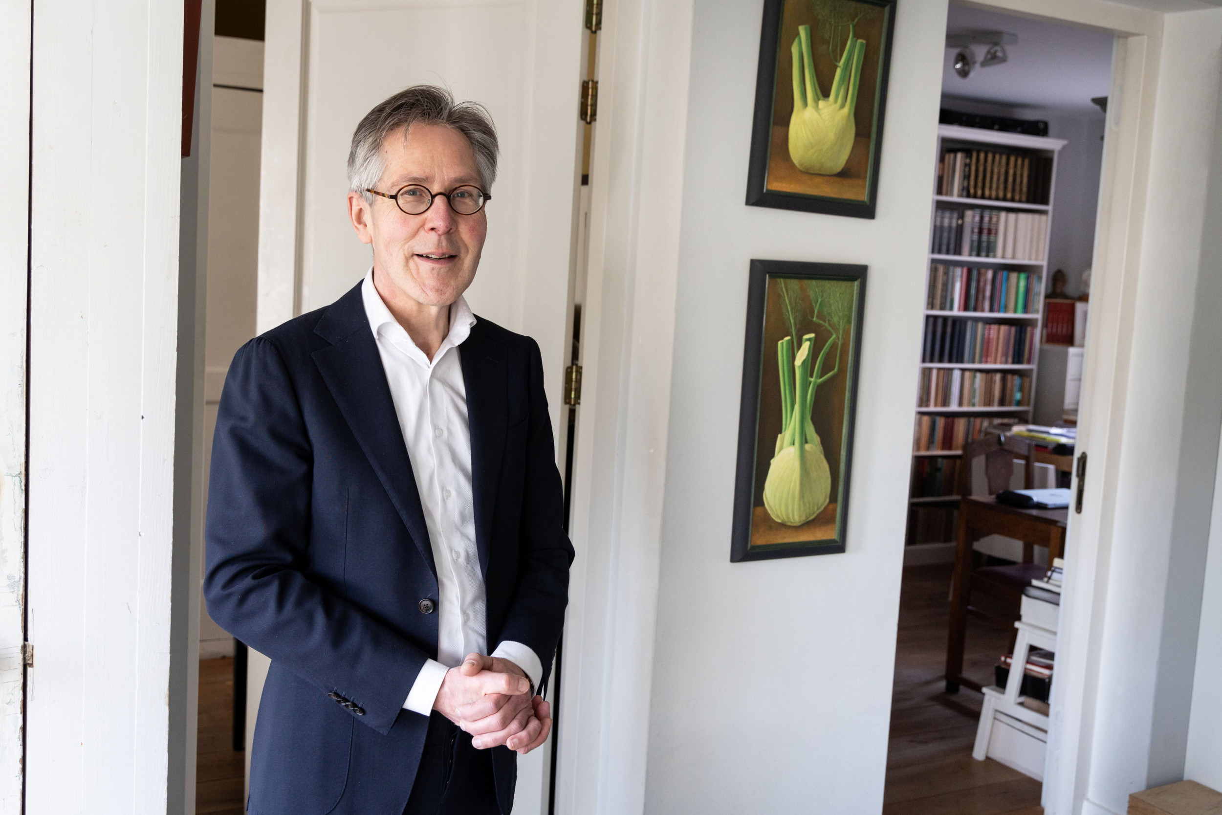 Man van middelbare leeftijd in donkerblauw kostuum in de deuropening van een kamer met boekenkast en schilderijen aan de wand.