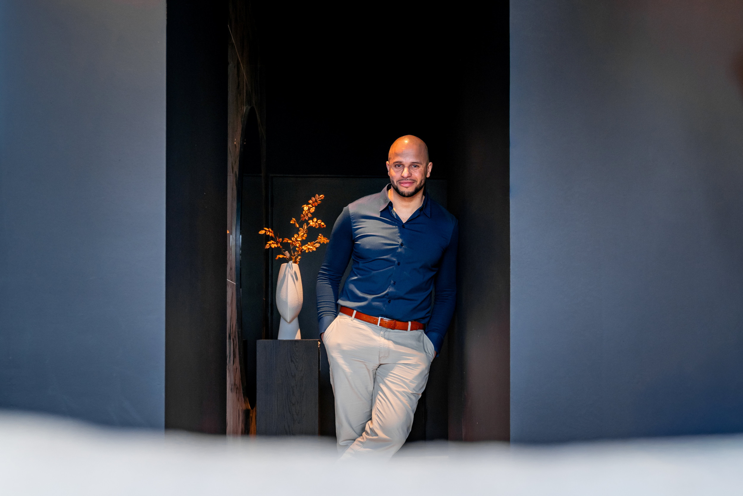 Man in blauw overhemd en lichte broek, leunt tegen een blauwe muur. Links van hem een vaas met een tak.