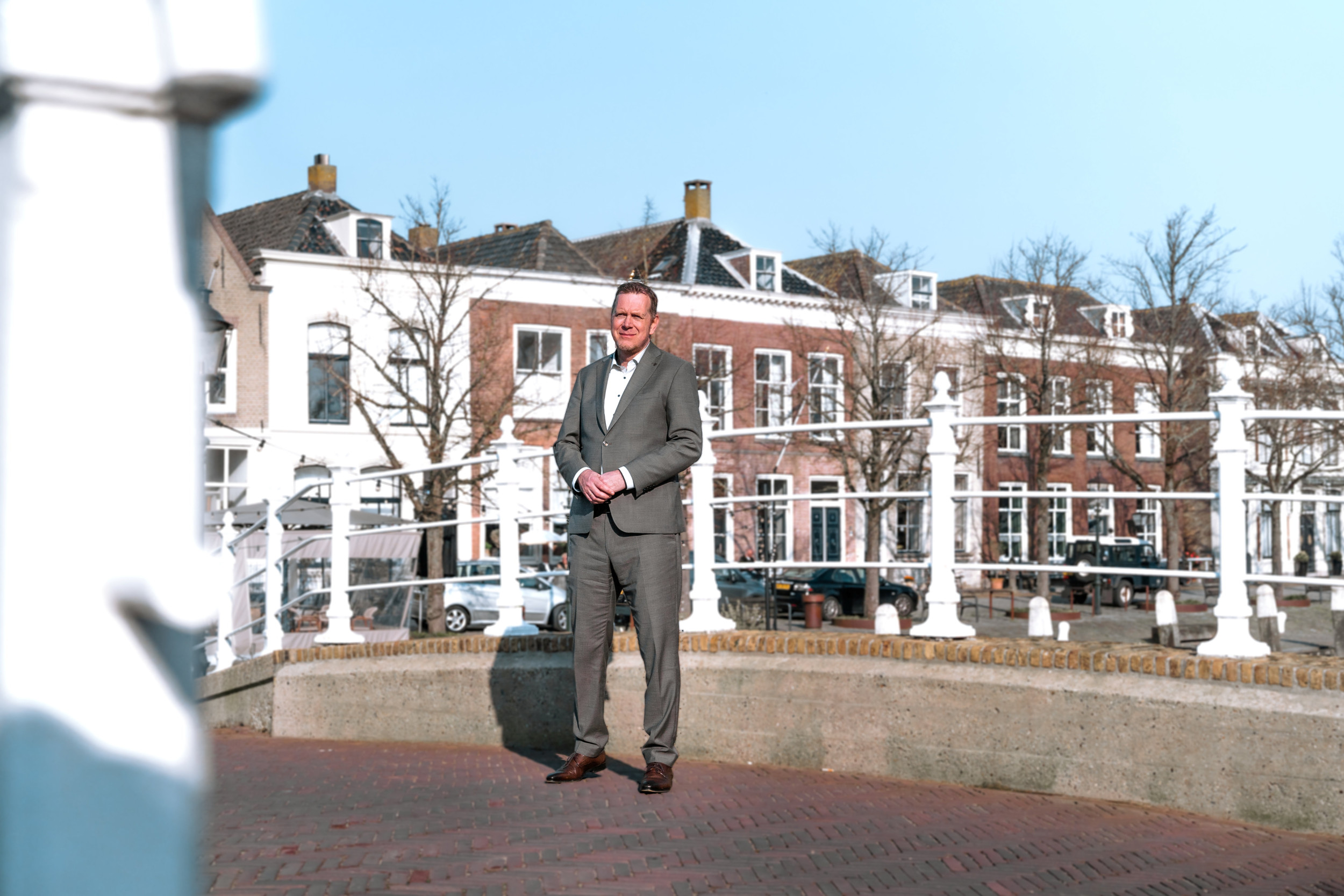 Man in grijs pak, staand op een brug, met op de achtergrond historische gevels.