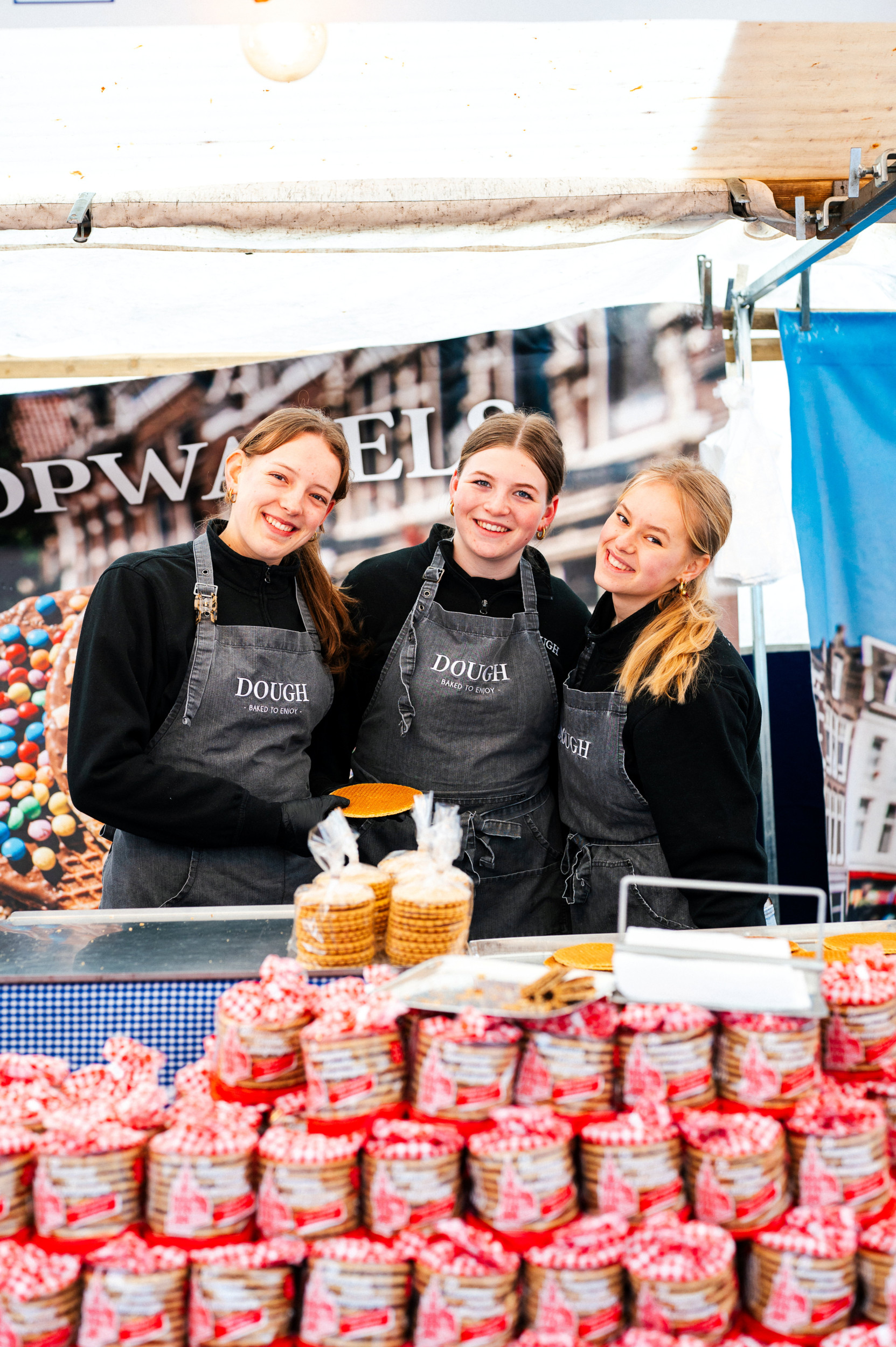 Drie jonge vrouwen in zwarte trui en grijze schort. Op de voorgrond pakjes met stroopkoeken.