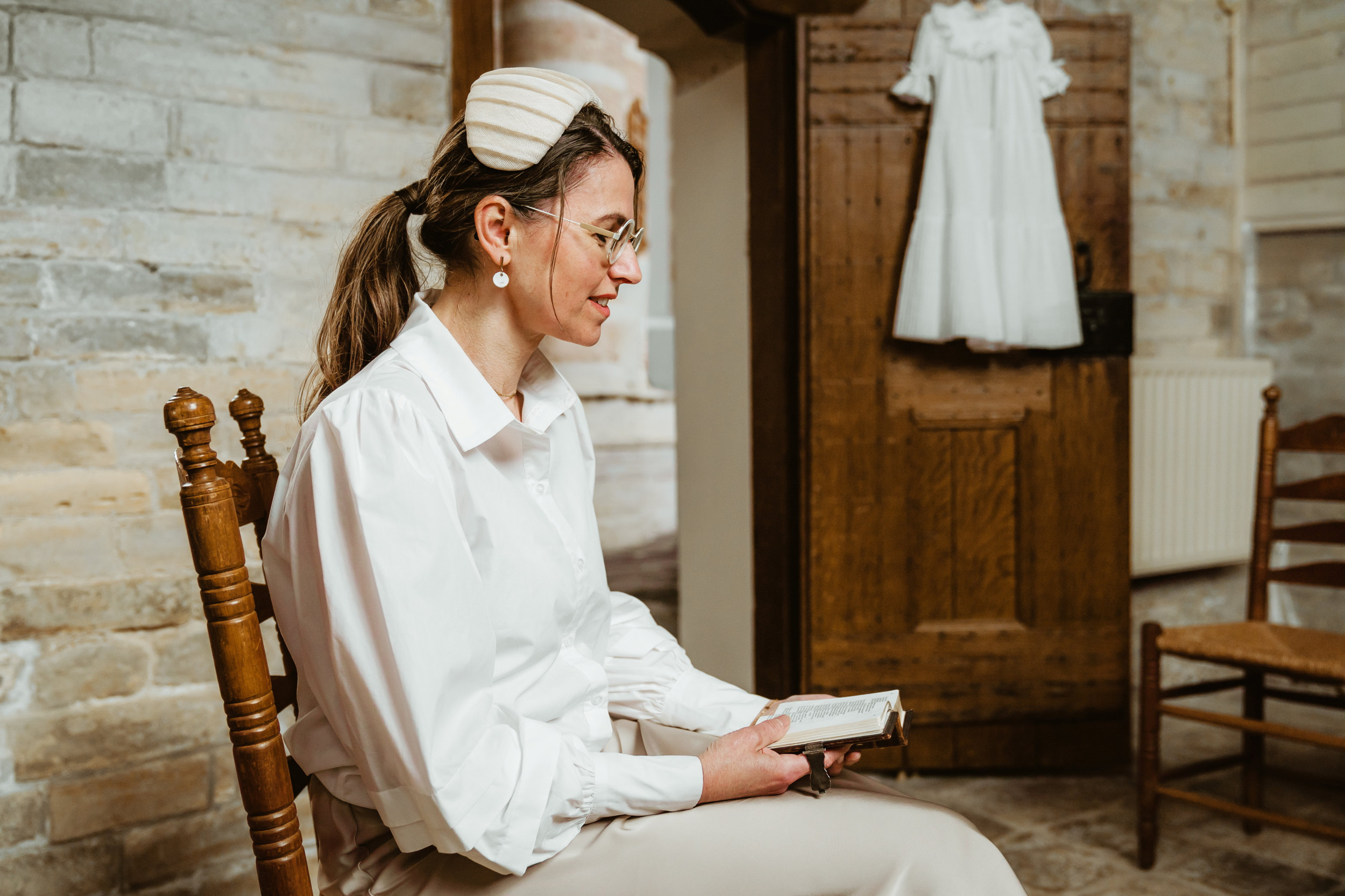 In het wit geklede vrouw met wit hoedje, zittend in een kerk waar ze uit een Bijbeltje leest. Aan een kast naast haar hangt een doopjurk.