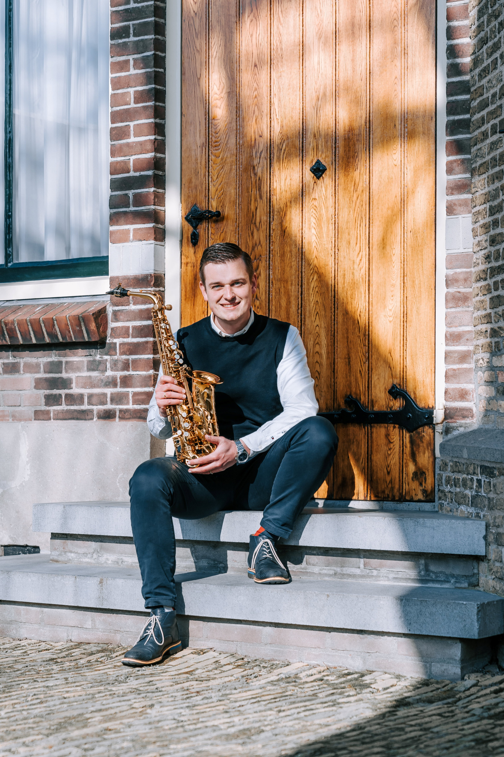 Jongeman in donkerblauwe spencer en licht overhemd, zittend op een stoepje, met een saxofoon in zijn handen.