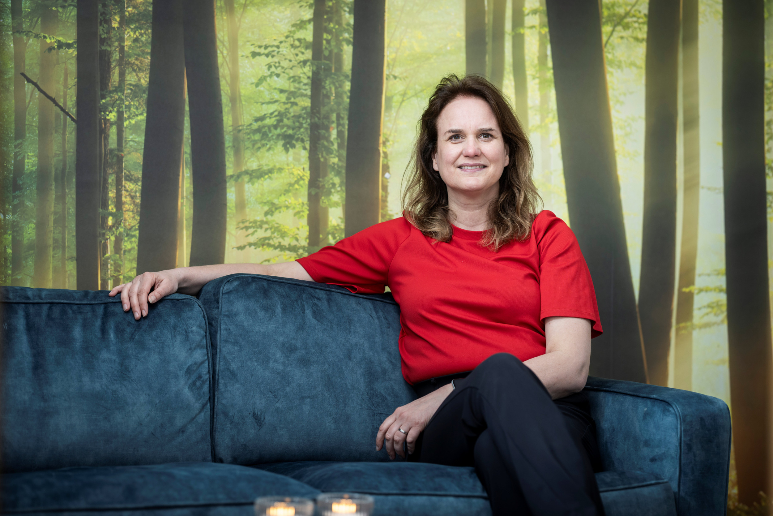 Donkerblonde vrouw, met een helderrood shirt aan op een blauwe bank. Op de achtergrond een wandvullende foto van een bos.