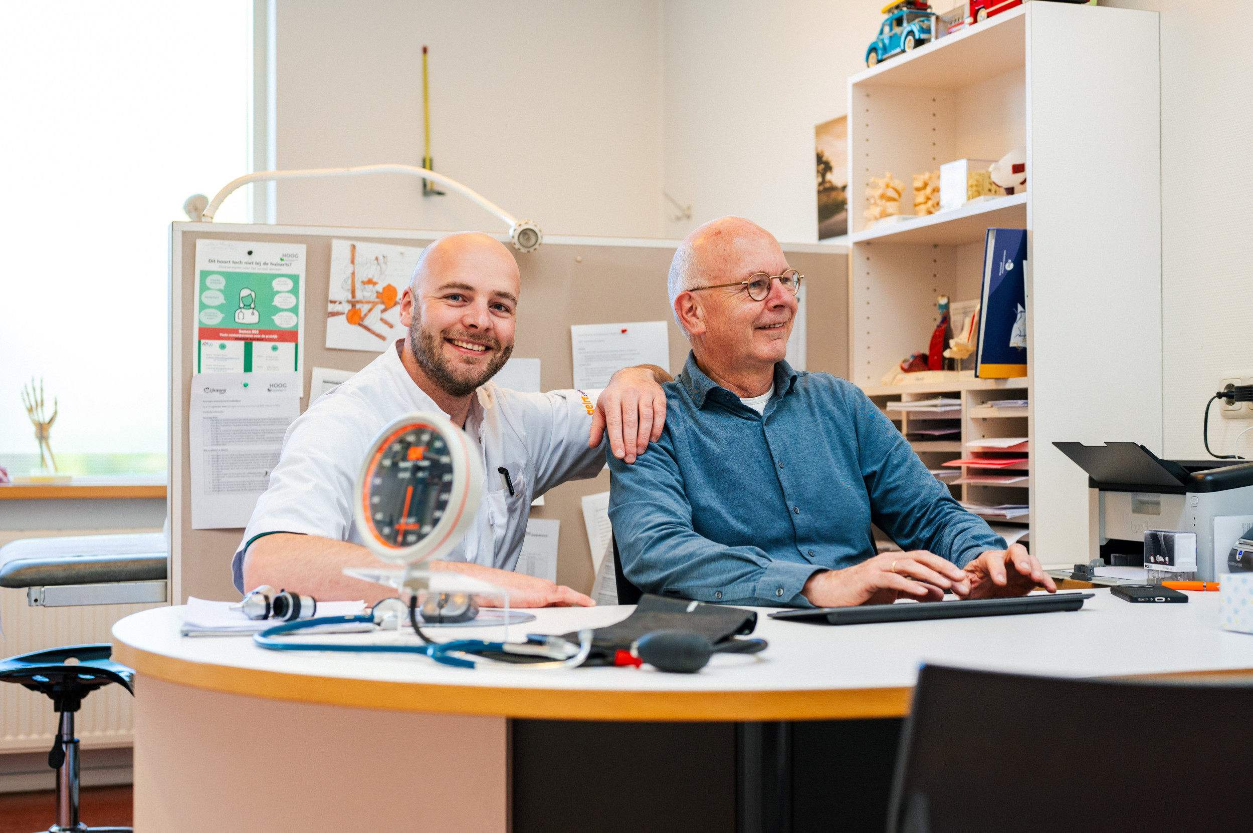 Jonge man in witte doktersjas naast oudere man met blauw overhemd aan een bureau.
