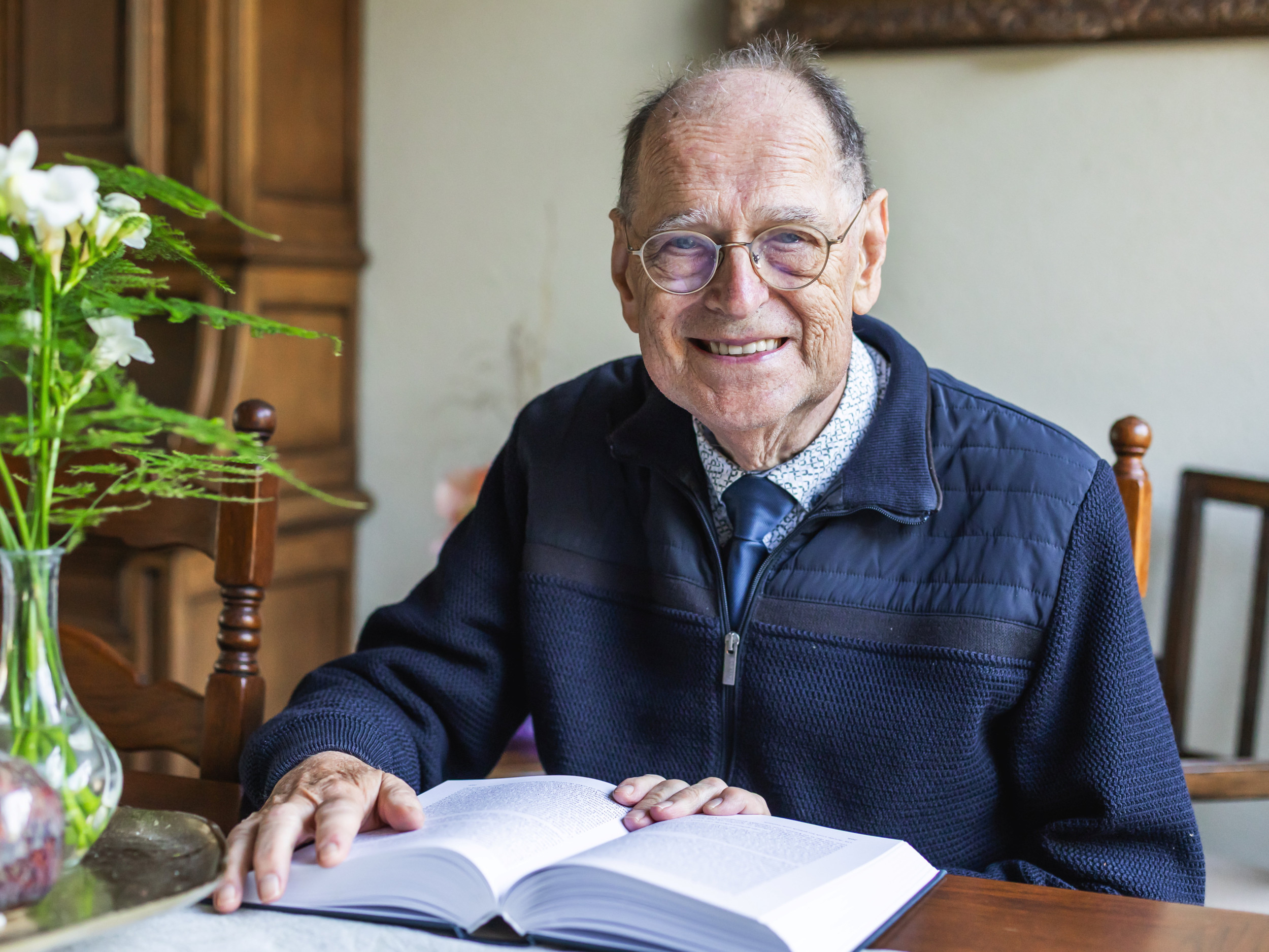 Oude man met een blauwe trui aan een tafel met een boek voor zich.