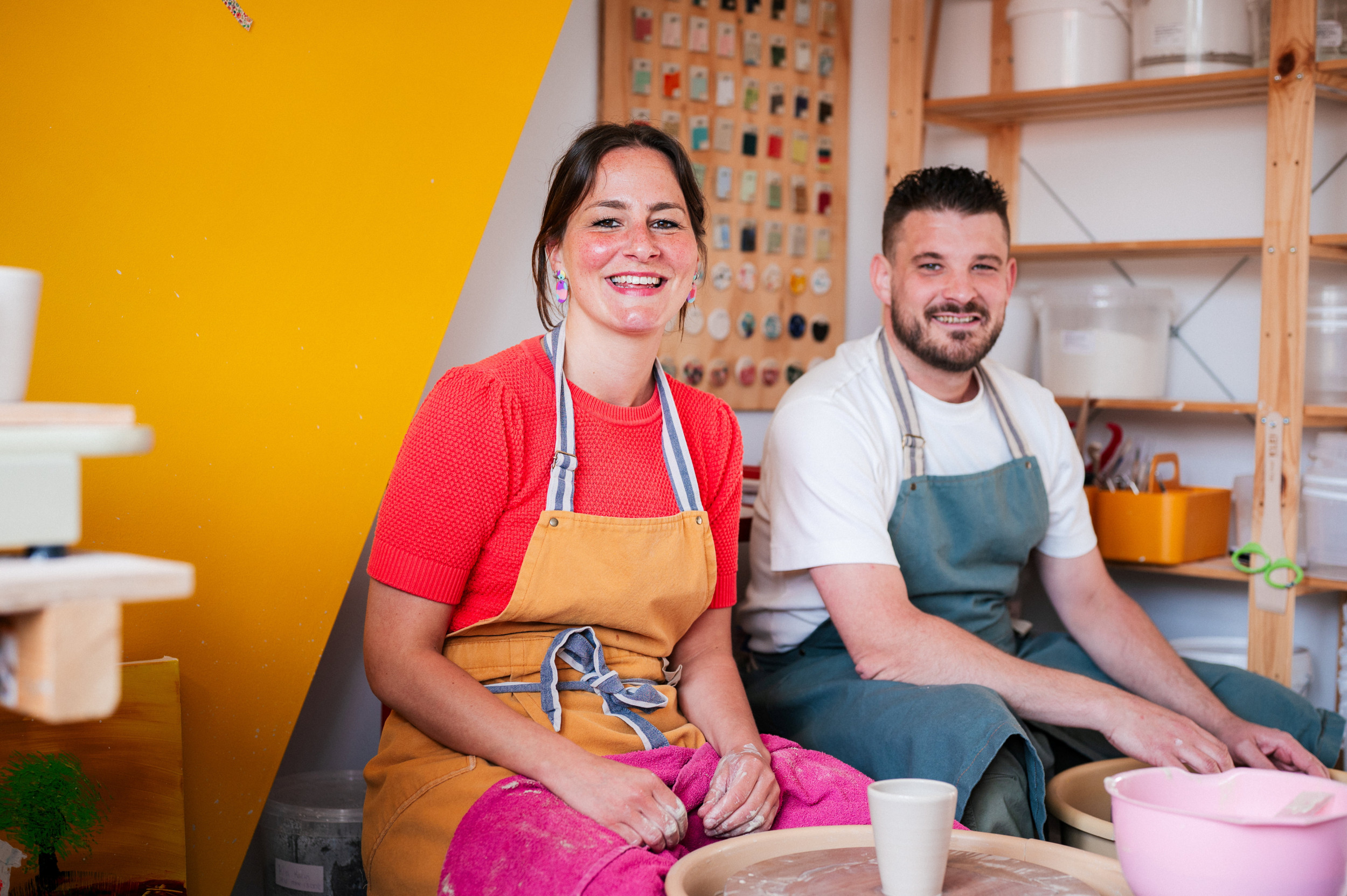 Links een lachende vrouw met donker haar en een oranje schort om, rechts een man met donker haar en een blauw short voor. Beiden zitten voor een pottenbakkersschijf.