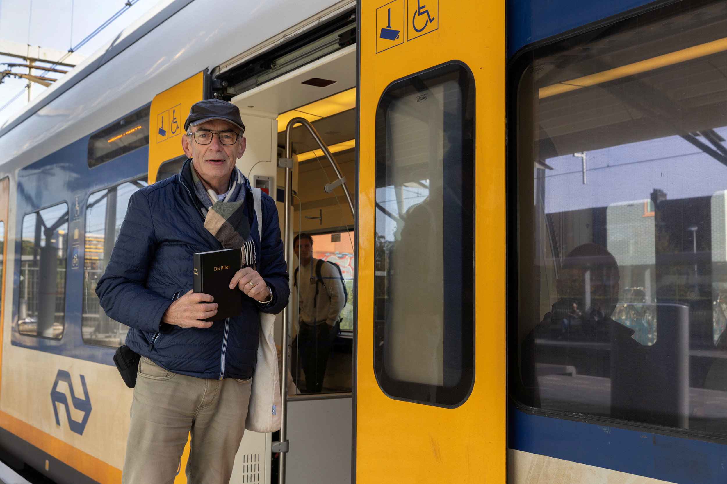 Man met bril op, beige broek en donkerblauwe jas en pet, met een zwarte Bijbel in zijn handen; staande voor de geopende deuren van een NS-trein. 