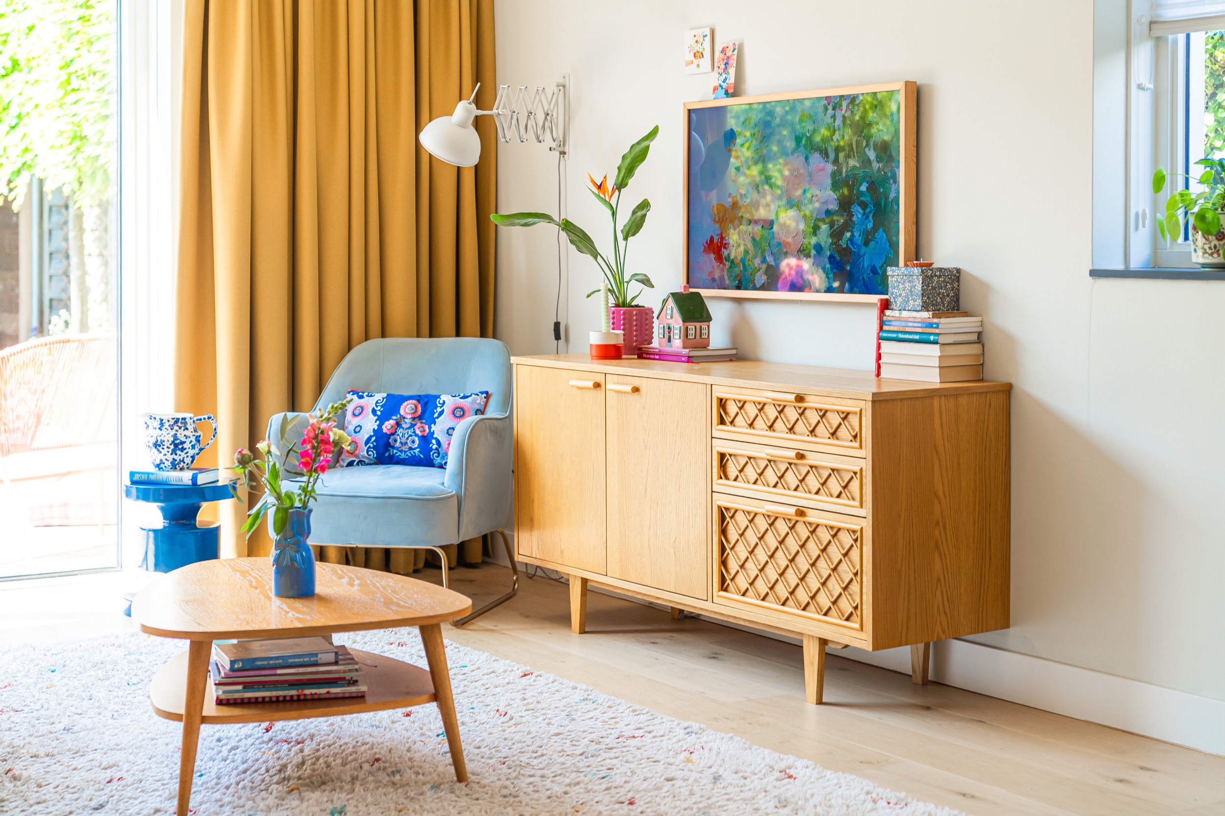 De woonkamer van Heidi Kuiper: een vintage kastje, een lichtblauwe leunstoel, gele gordijnen en kleurige accessoires.
