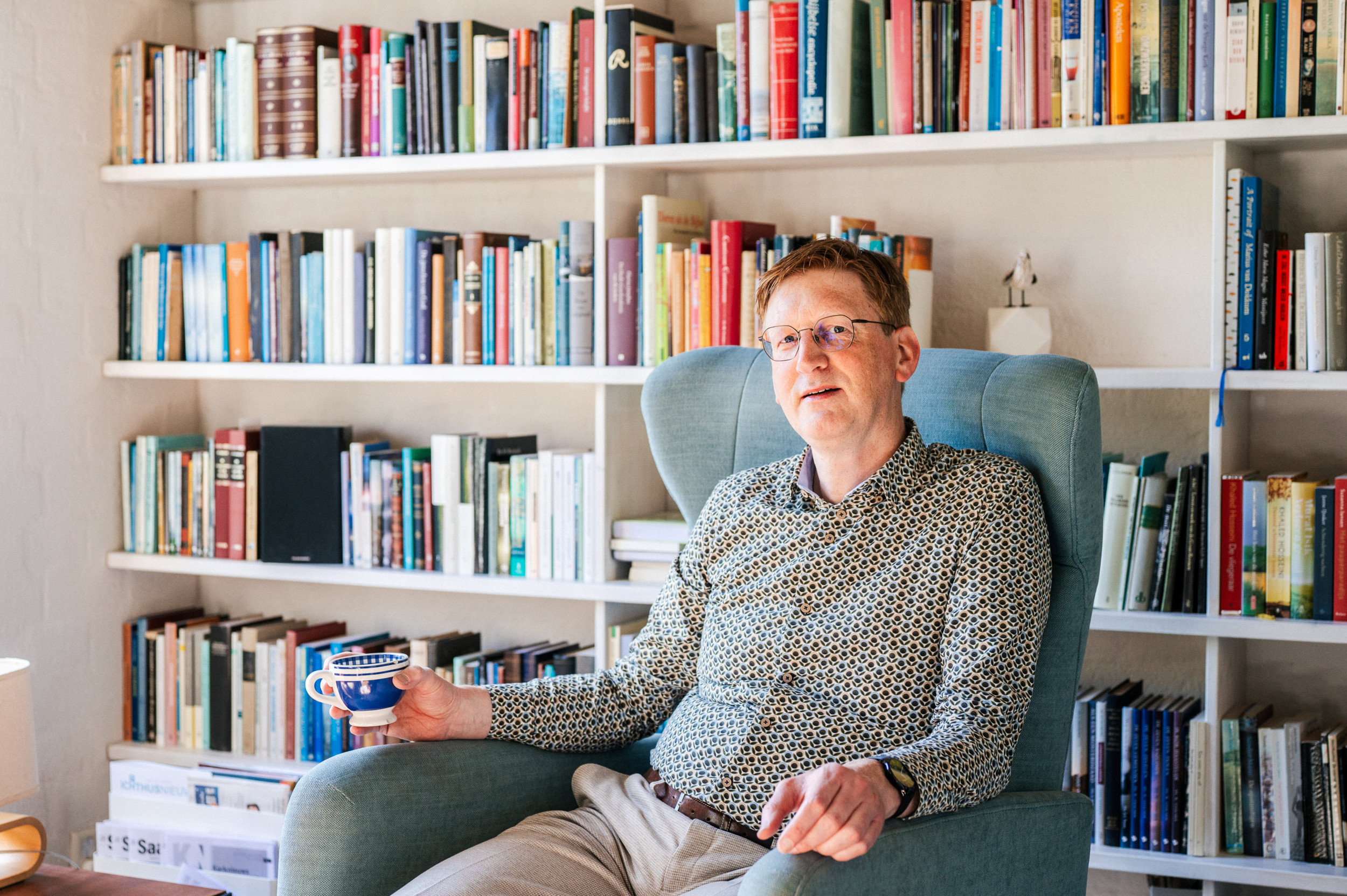 Man van middelbare leeftijd met rood haar voor een boekenkast met een koffiebeker in de hand.