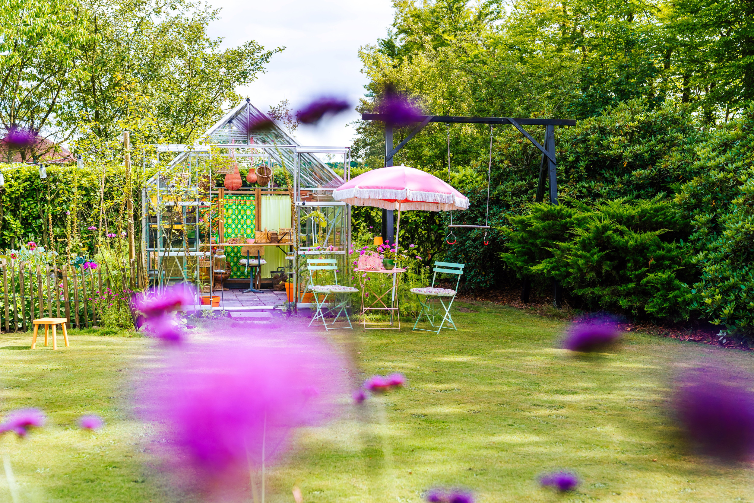 Groene, fleurige tuin met kas, schommel en parasol.