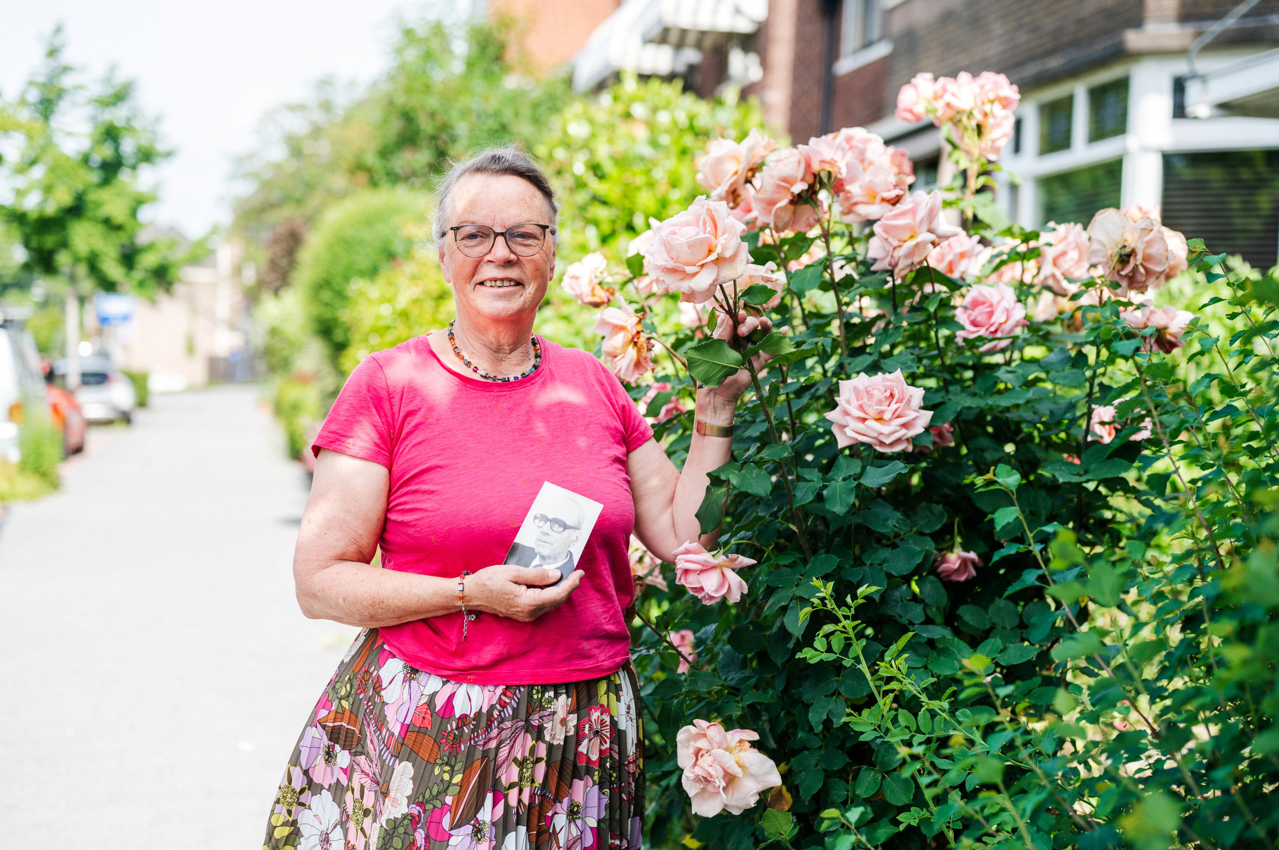 Vrouw van middelbare leeftijd met roze shirt en gebloemde jurk voor een rozenstruik, in haar rechterhand het zwartwitportret van een oudere man.