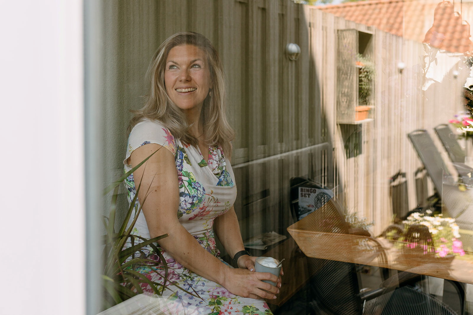 Blonde vrouw met een gebloemde jurk aan en een kop cappuccino in haar handen. Ze kijkt met een lach door een raam naar buiten.