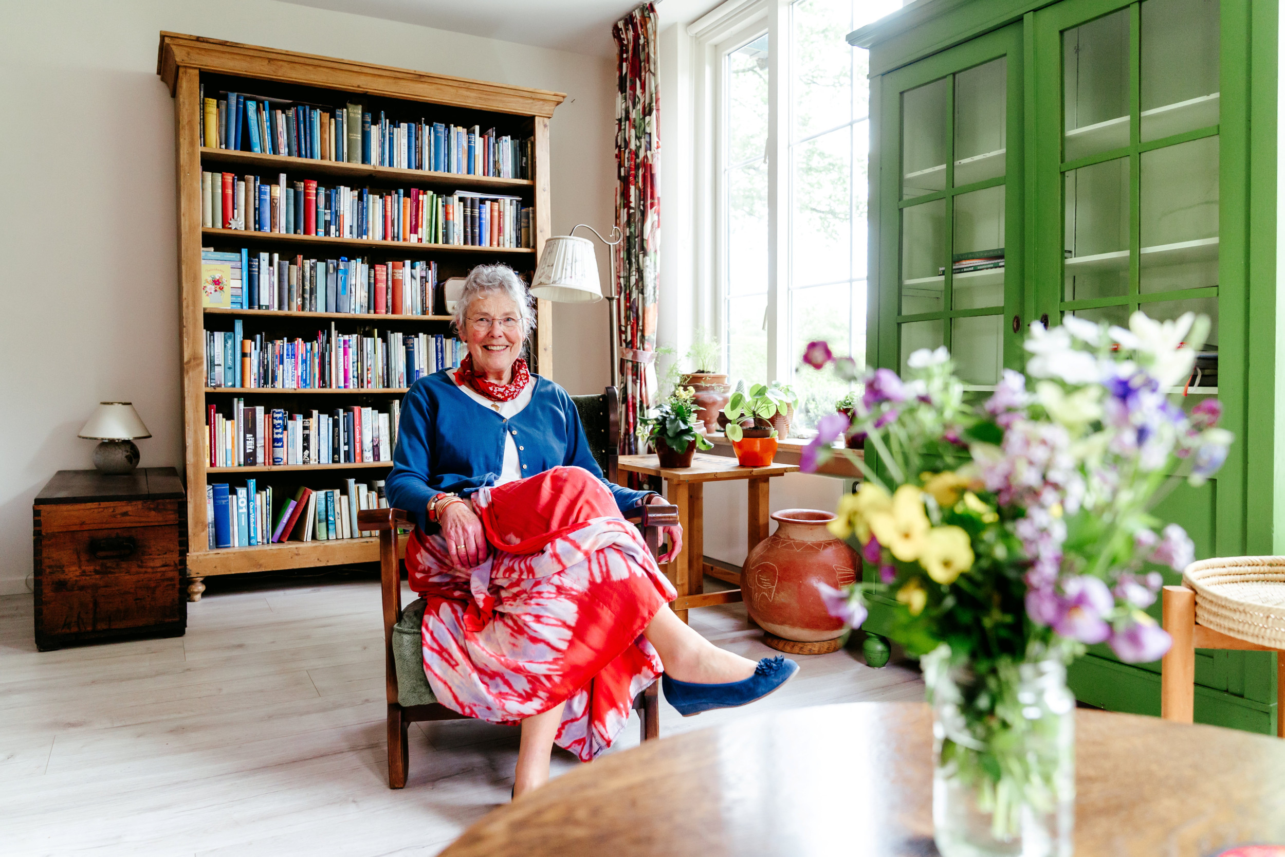 Vrouw met grijs haar, rode rok en blauw vestje, zittend op een stoel in een woonkamer.