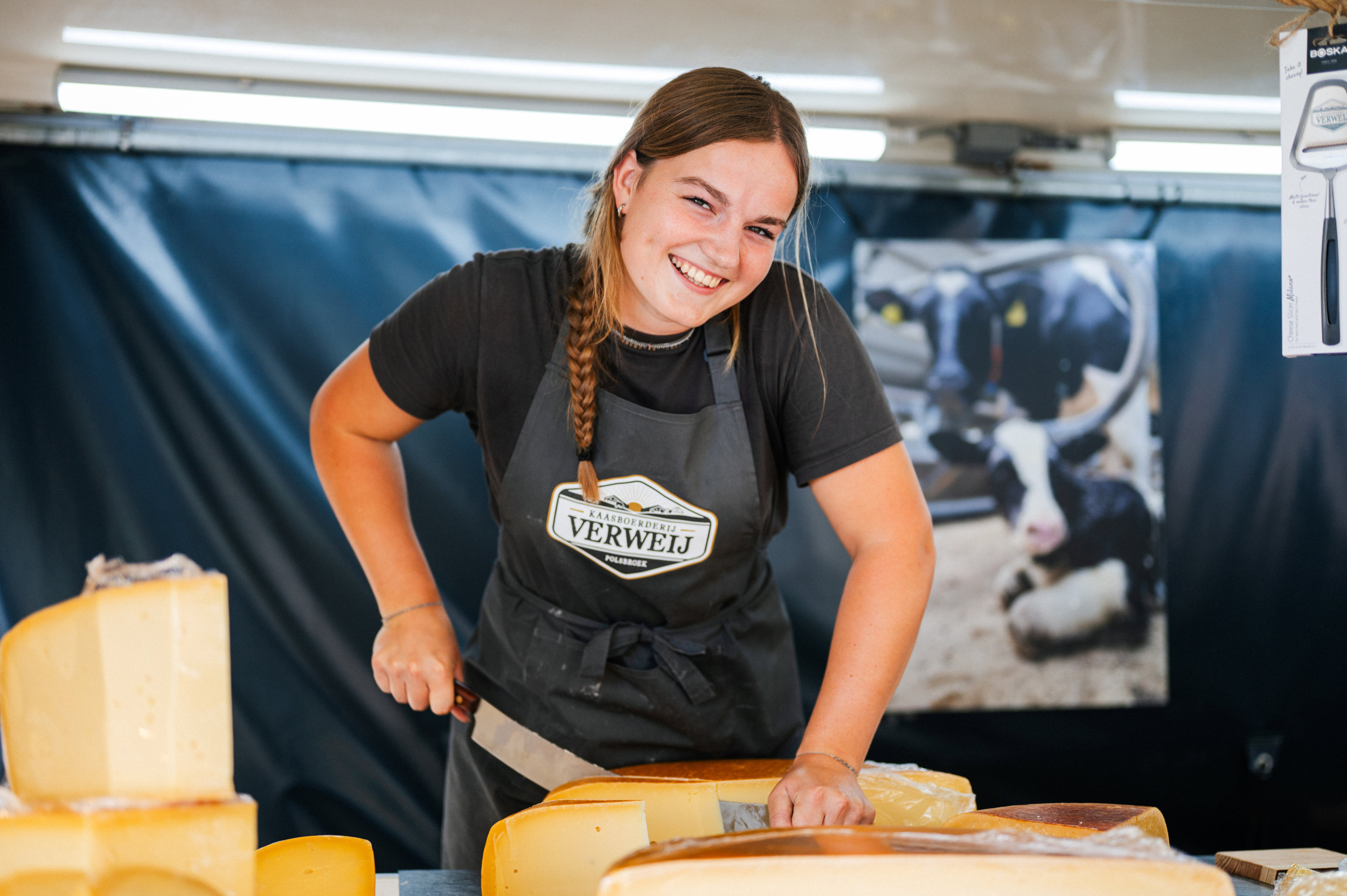 Jonge vrouw met zwart schort en zwart shirt snijdt met een groot kaasmes een stuk kaas af. Ze kijkt met een lach in de lens en heeft een lange vlecht in haar donkerblonde haar.