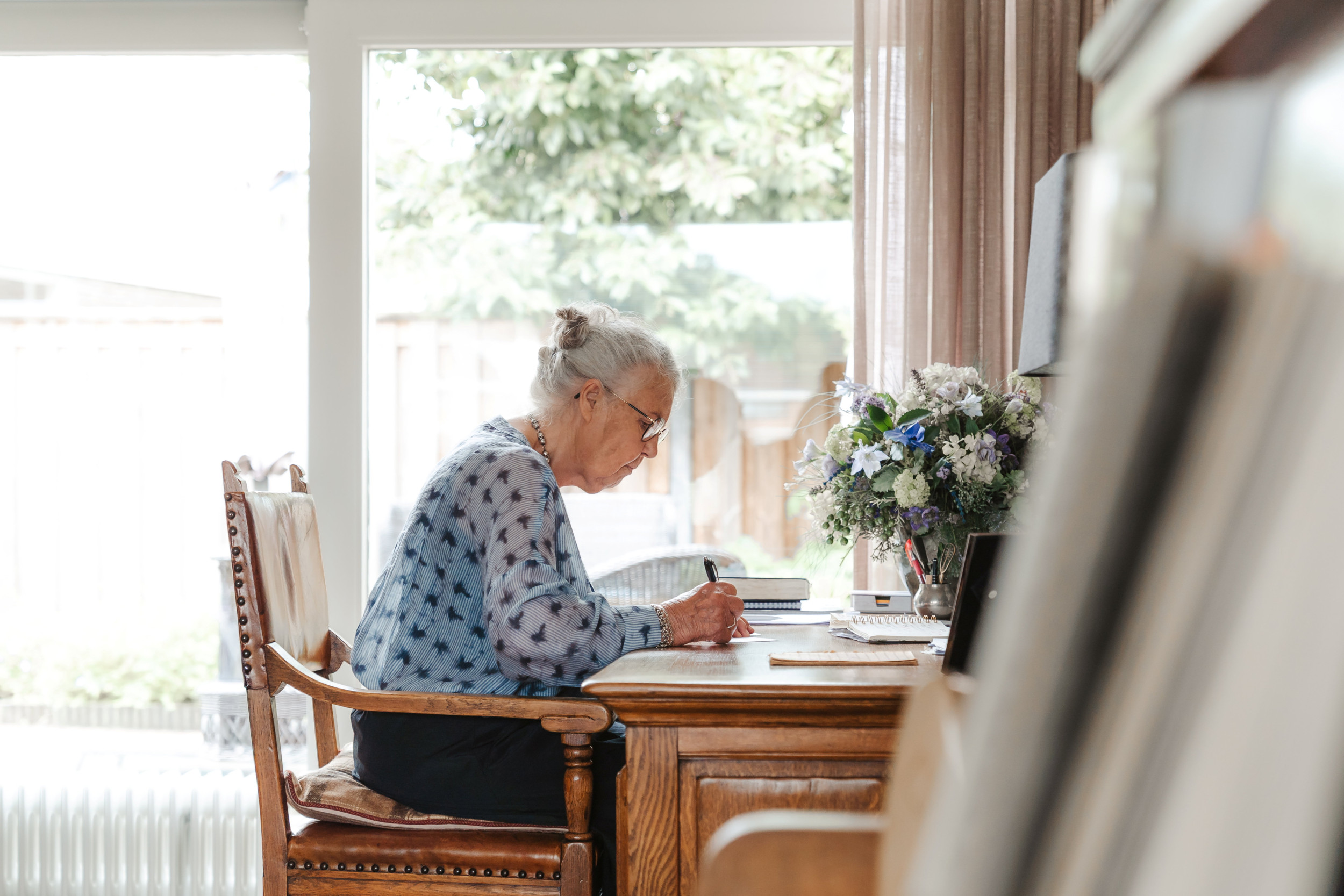 Oudere vrouw met grijs haar achter een bureau, terwijl ze iets opschrijft.