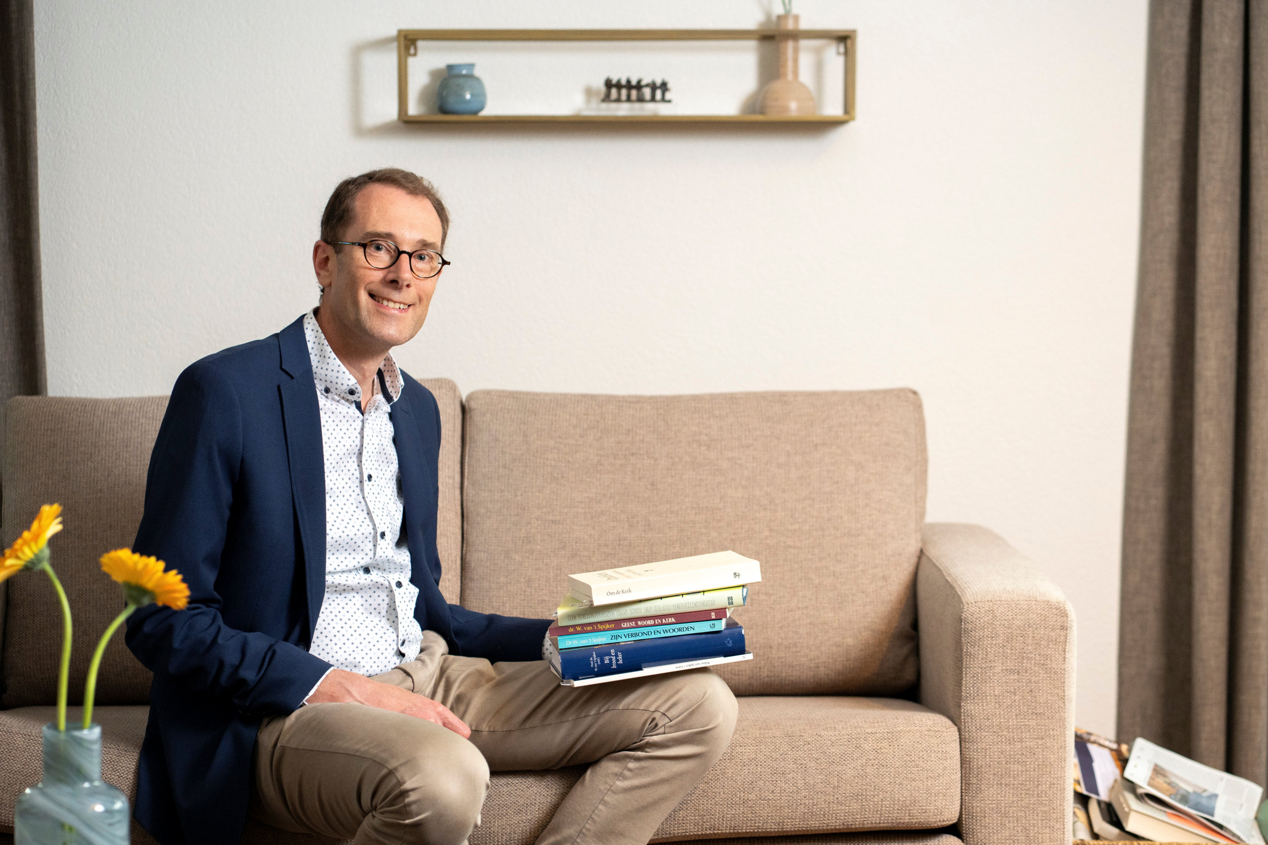 Man met lichte broek en blauw jasje, zittend op een bank met op zijn linkerbeen een stapel boeken.