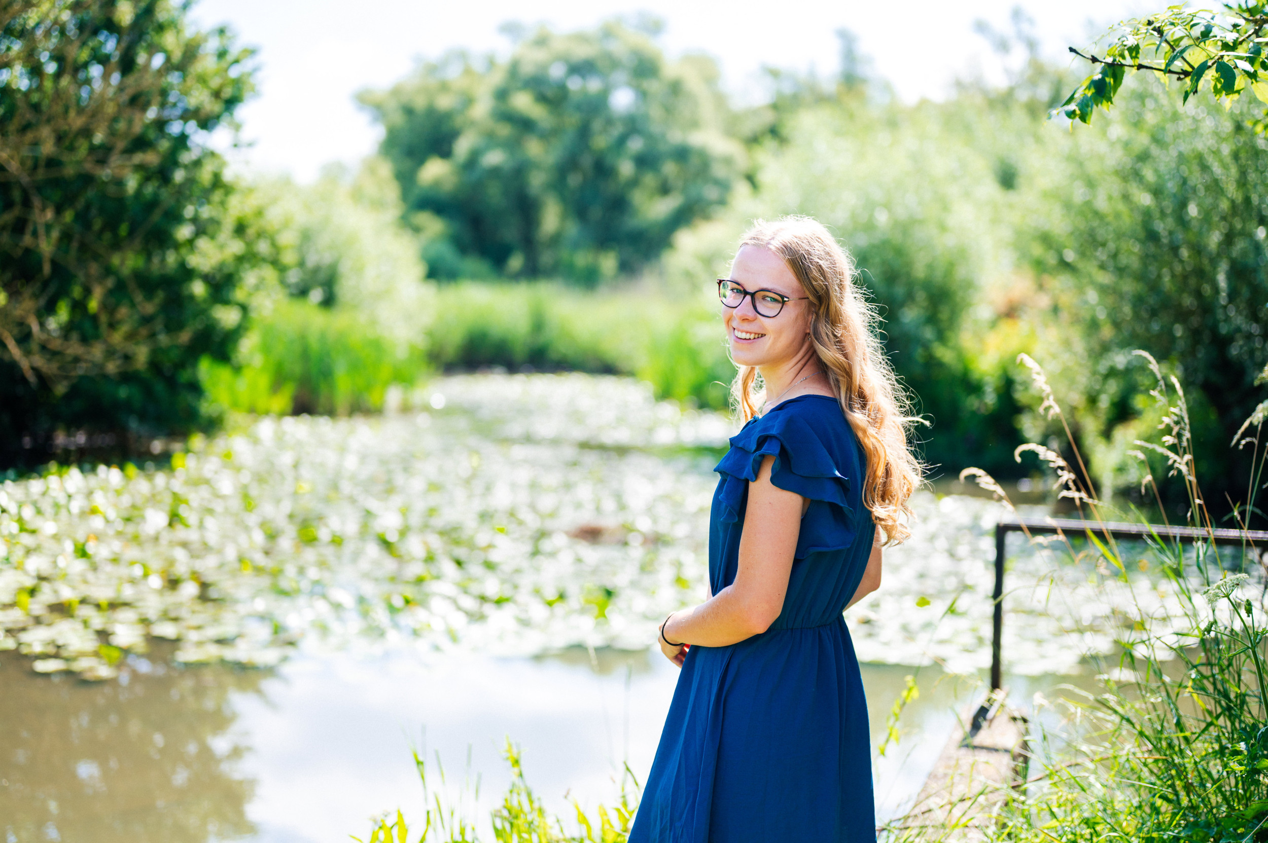 Jonge vrouw met blauwe jurk, lang, blond haar en een bril met donker montuur op. Op de achtergrond een vijver met waterlelies.