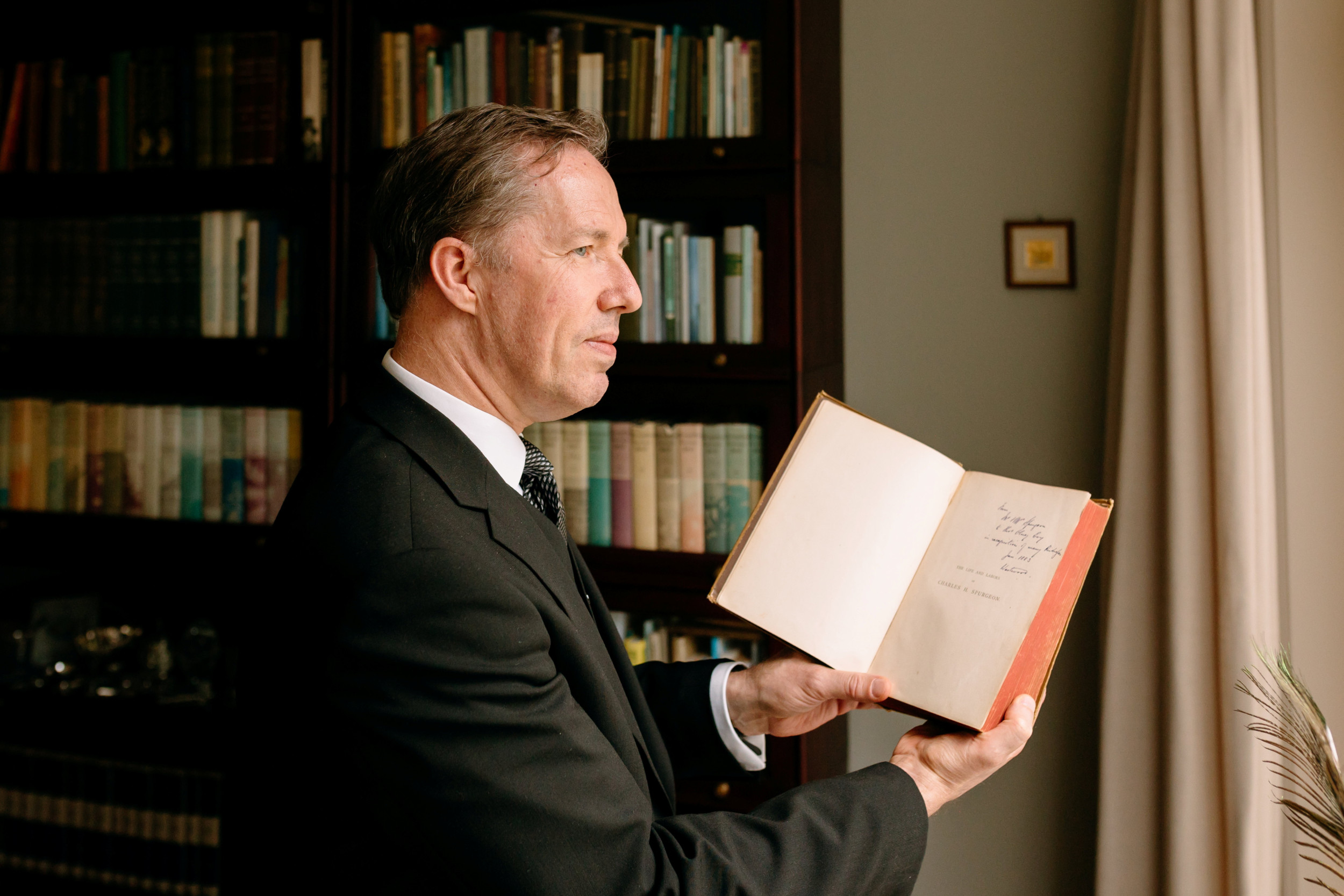 Man in donker pak voor een boekenkast met een boek in de hand.
